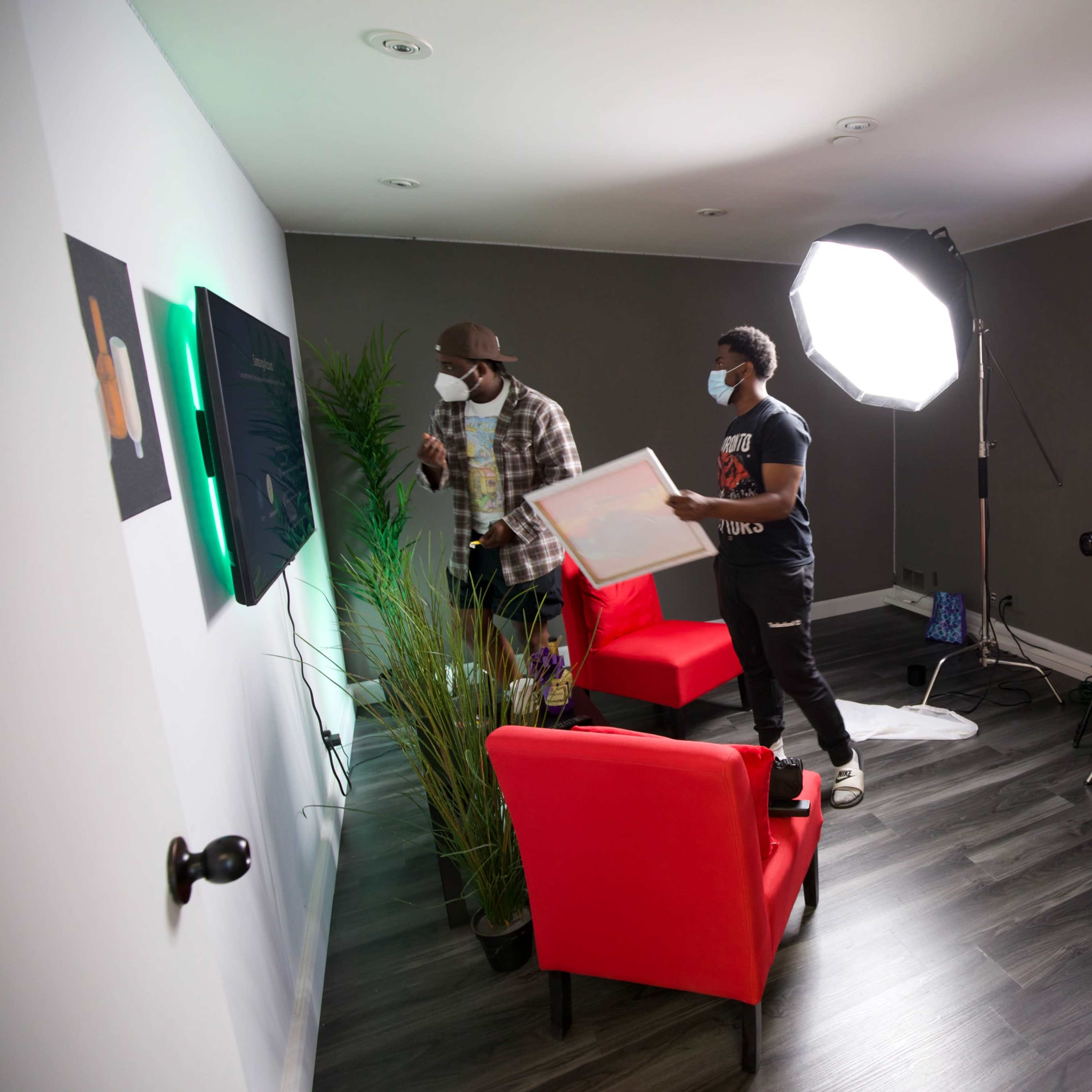 Three individuals set up a small video production scene with red chairs and lighting equipment in a dimly lit room.