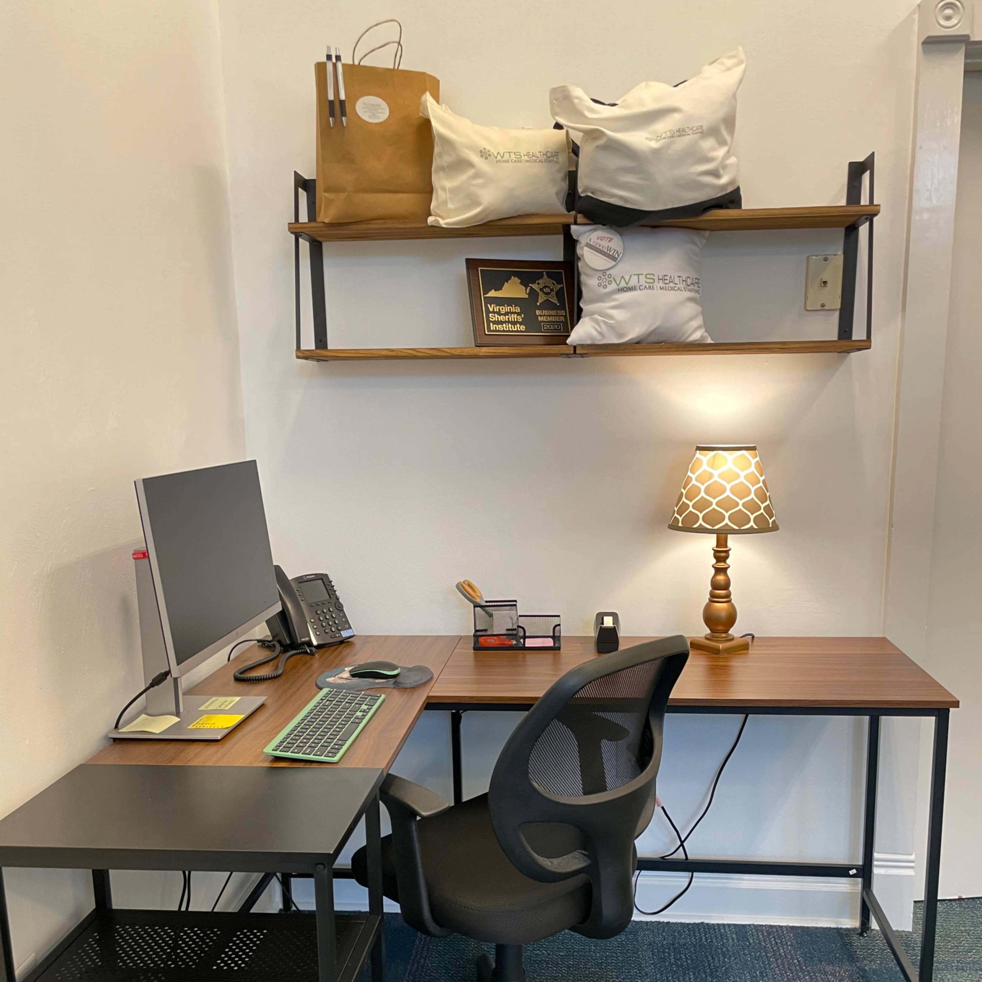 A workspace featuring a desk with a computer, a chair, a lamp, and shelves above with bags and decorative items.