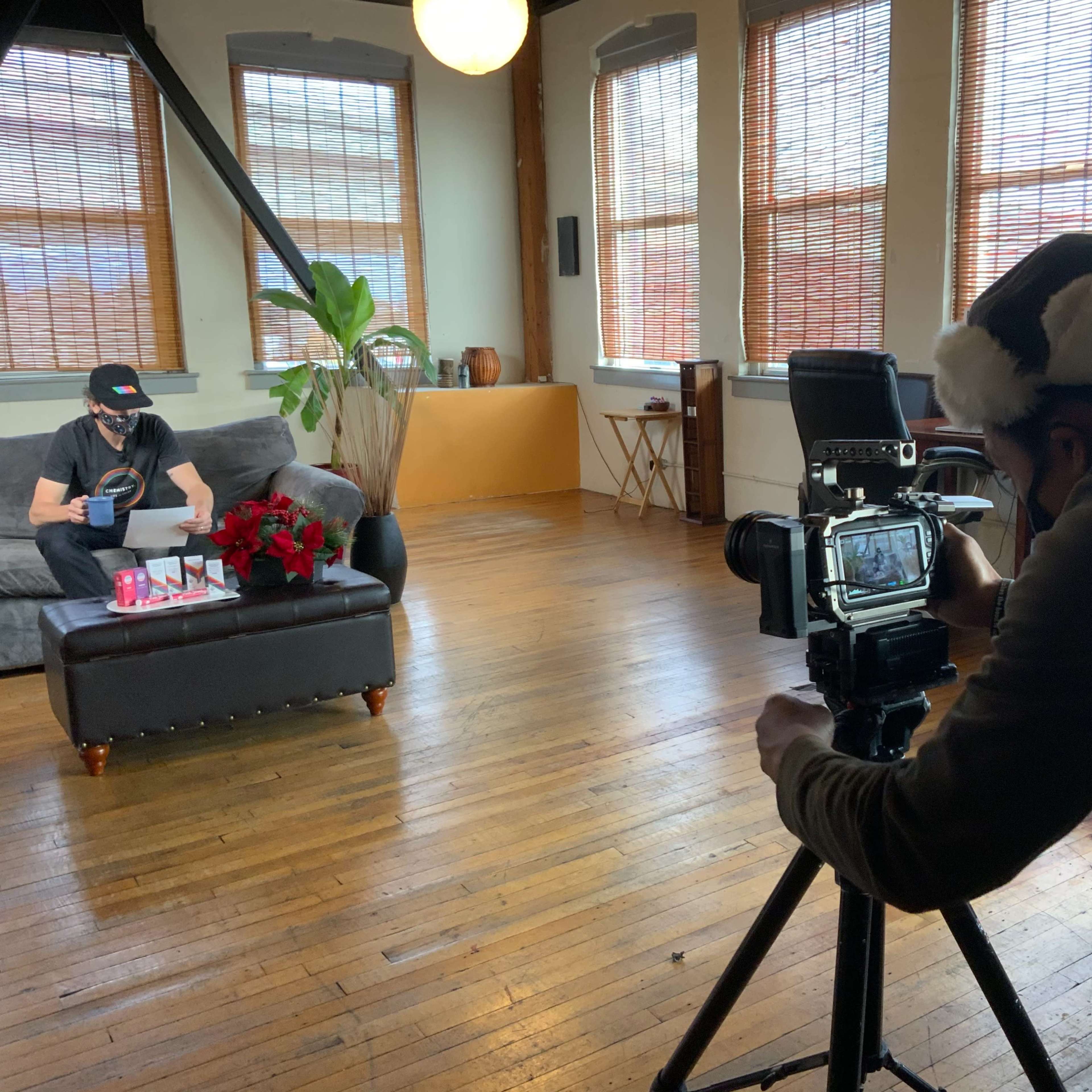 A person is filming another individual sitting on a couch with a camera and tripod in a sunlit room decorated with plants and wooden furnishings.