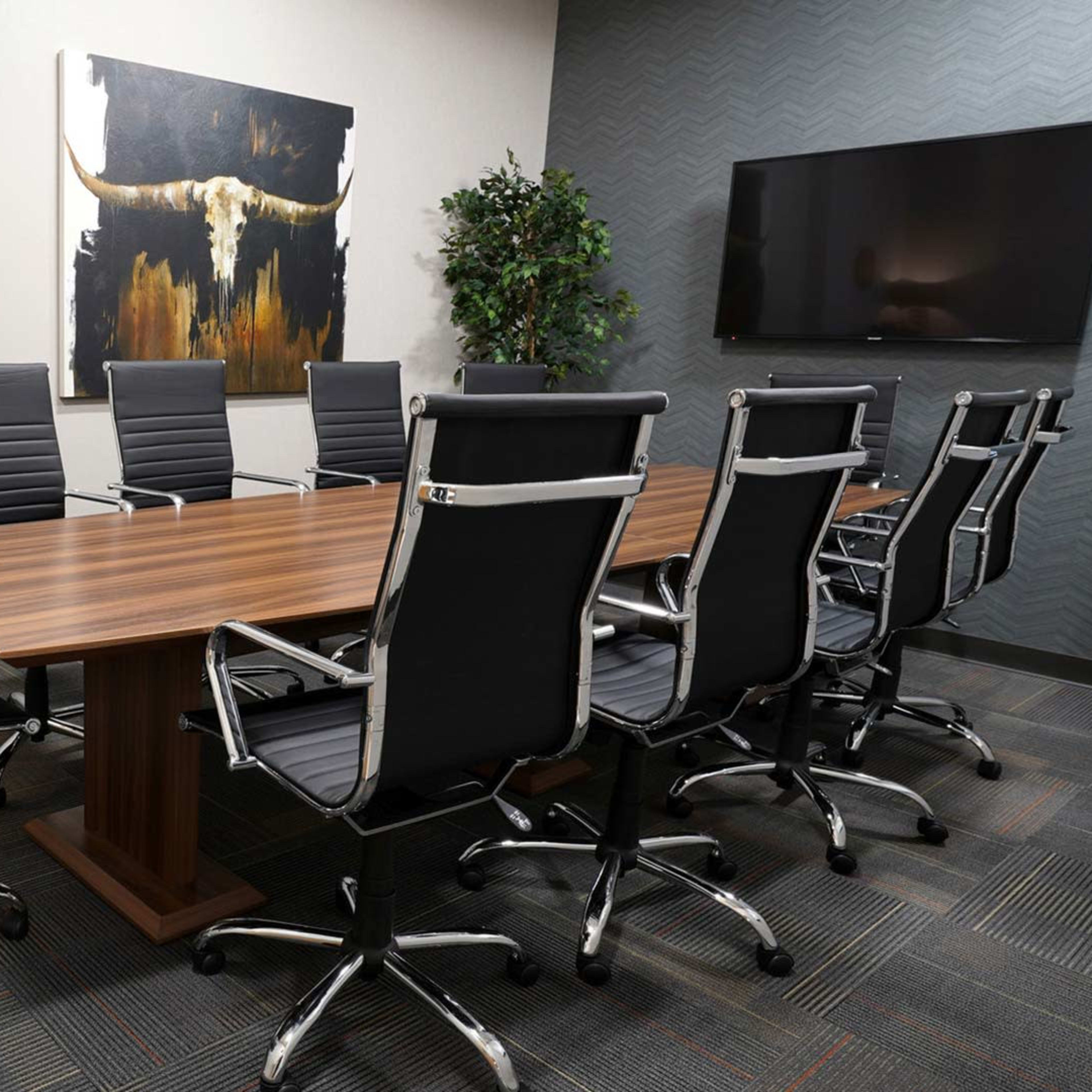 A modern conference room features a long wooden table surrounded by black ergonomic chairs, with a large television and decorative artwork on the walls.