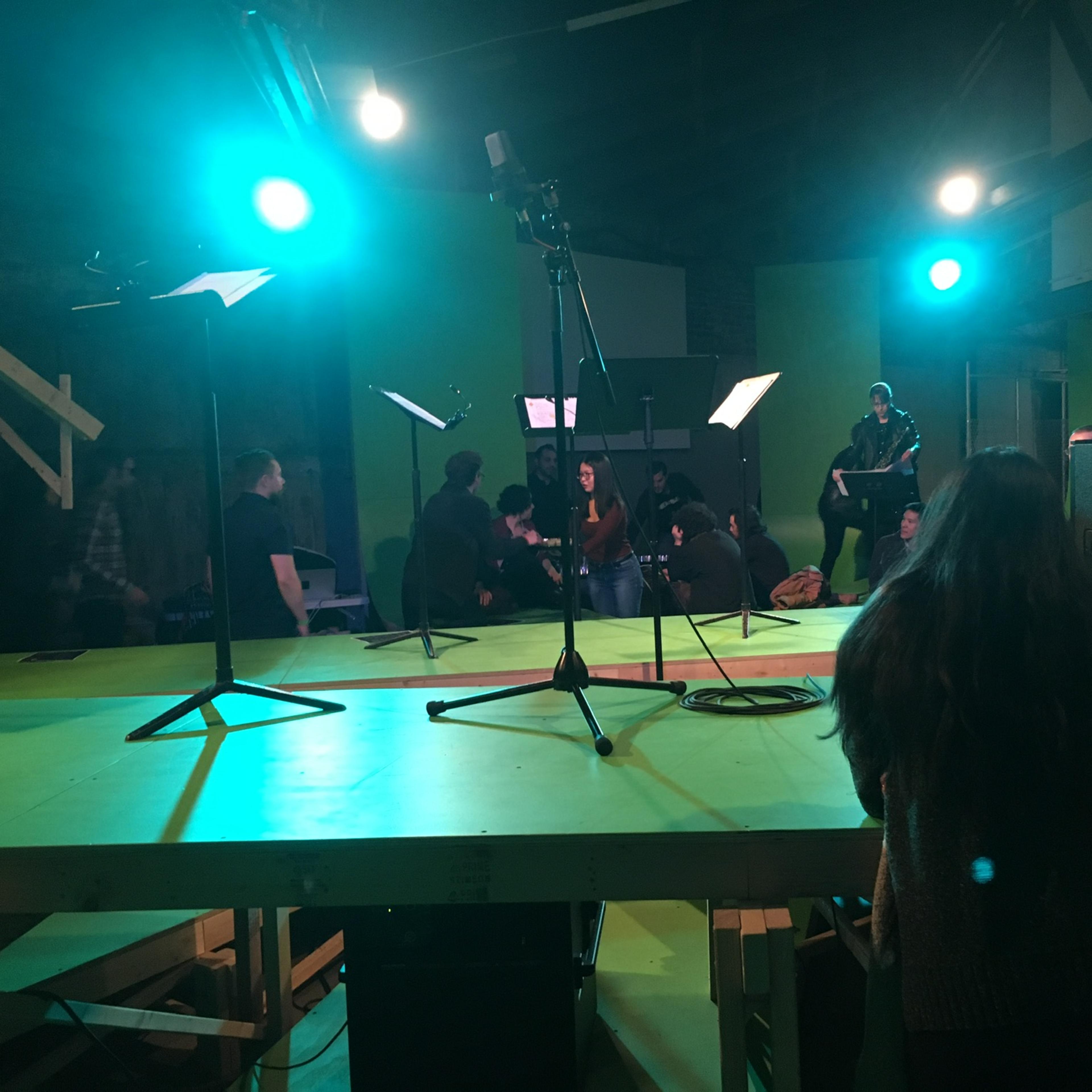 A group of people is gathered on a green stage with microphones and music stands in a well-lit room.