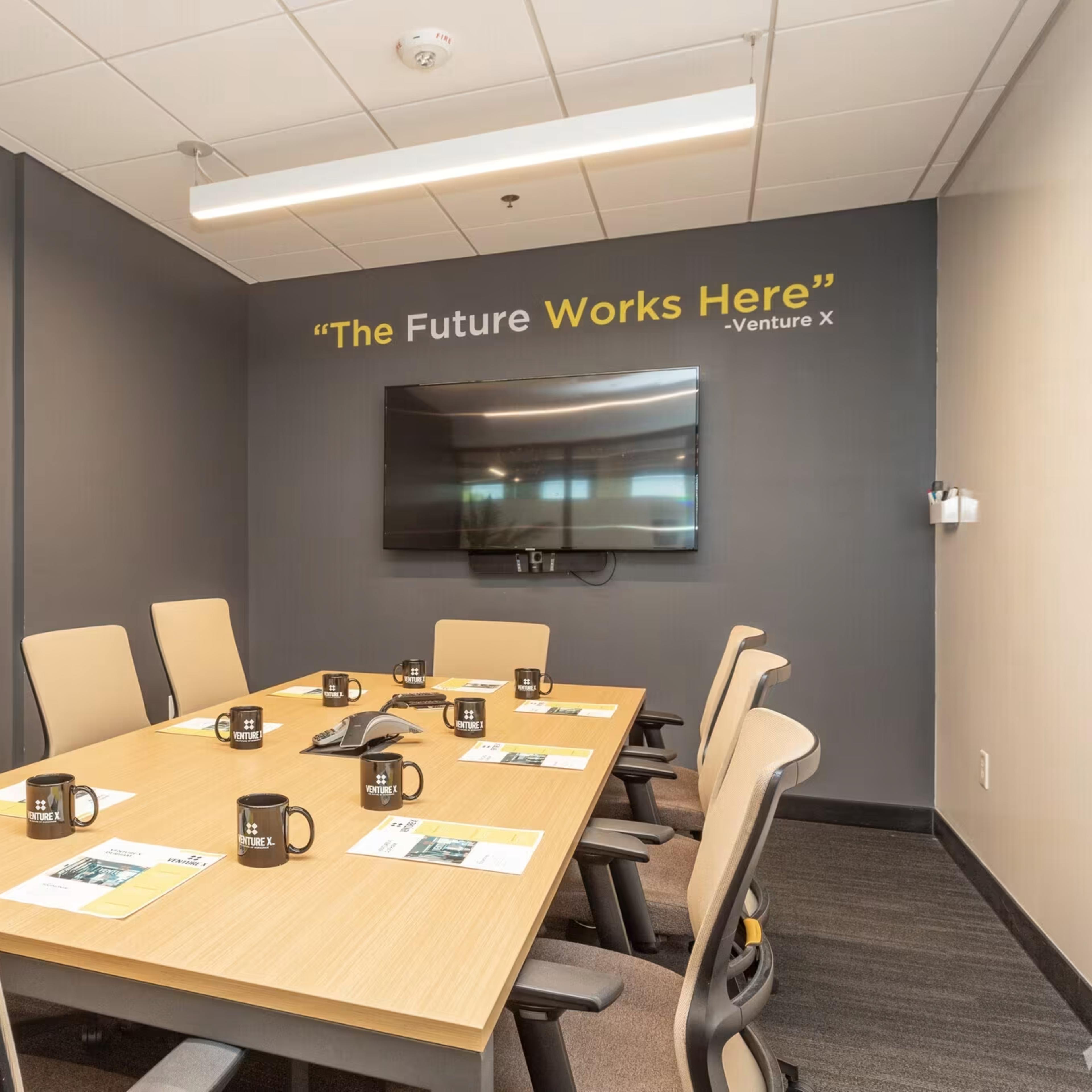 A conference room features a long table with six chairs, a large wall-mounted television, and a motivational quote above.