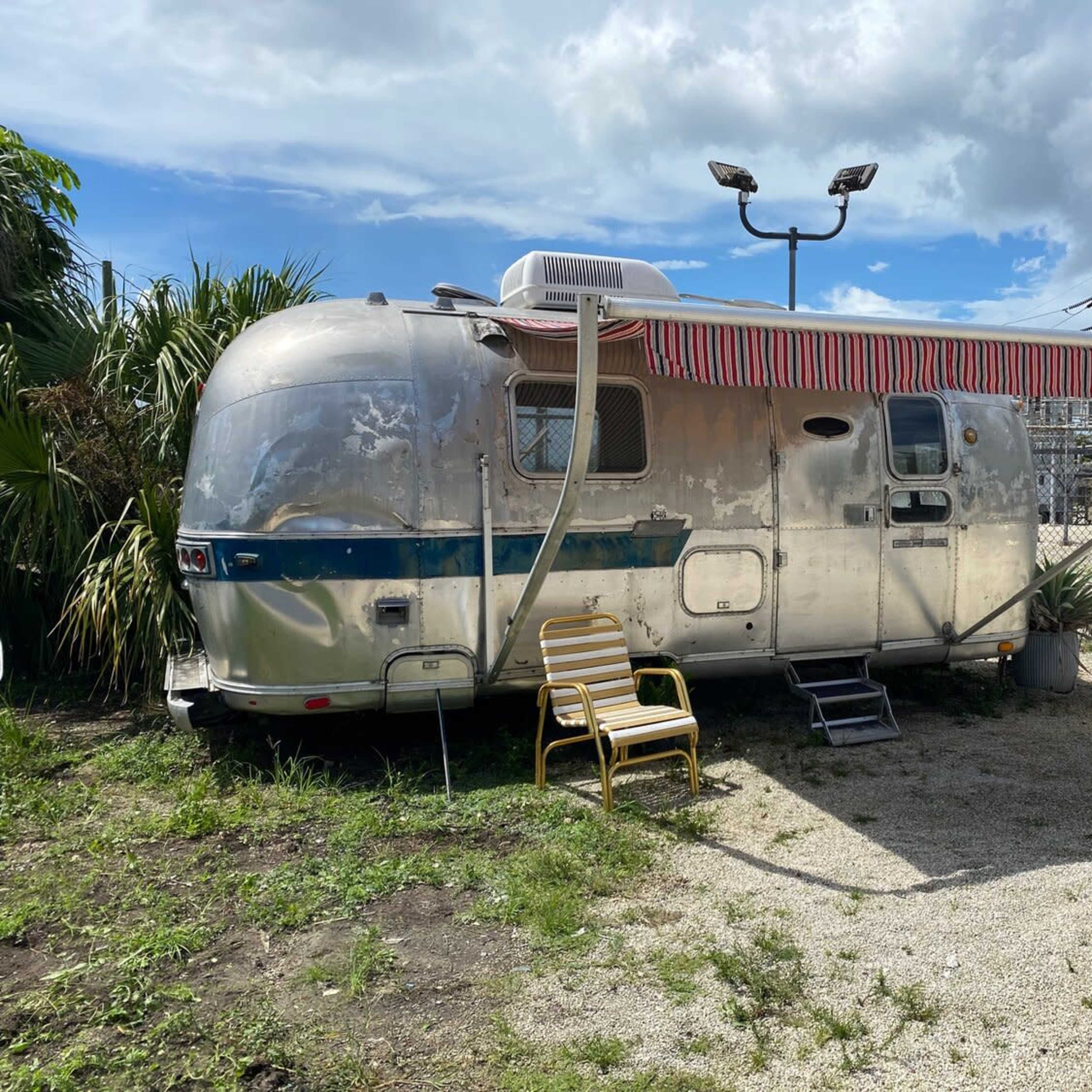 An aluminum Airstream trailer is parked next to a fence, with two yellow chairs positioned outside and overhead power lines in the background.
