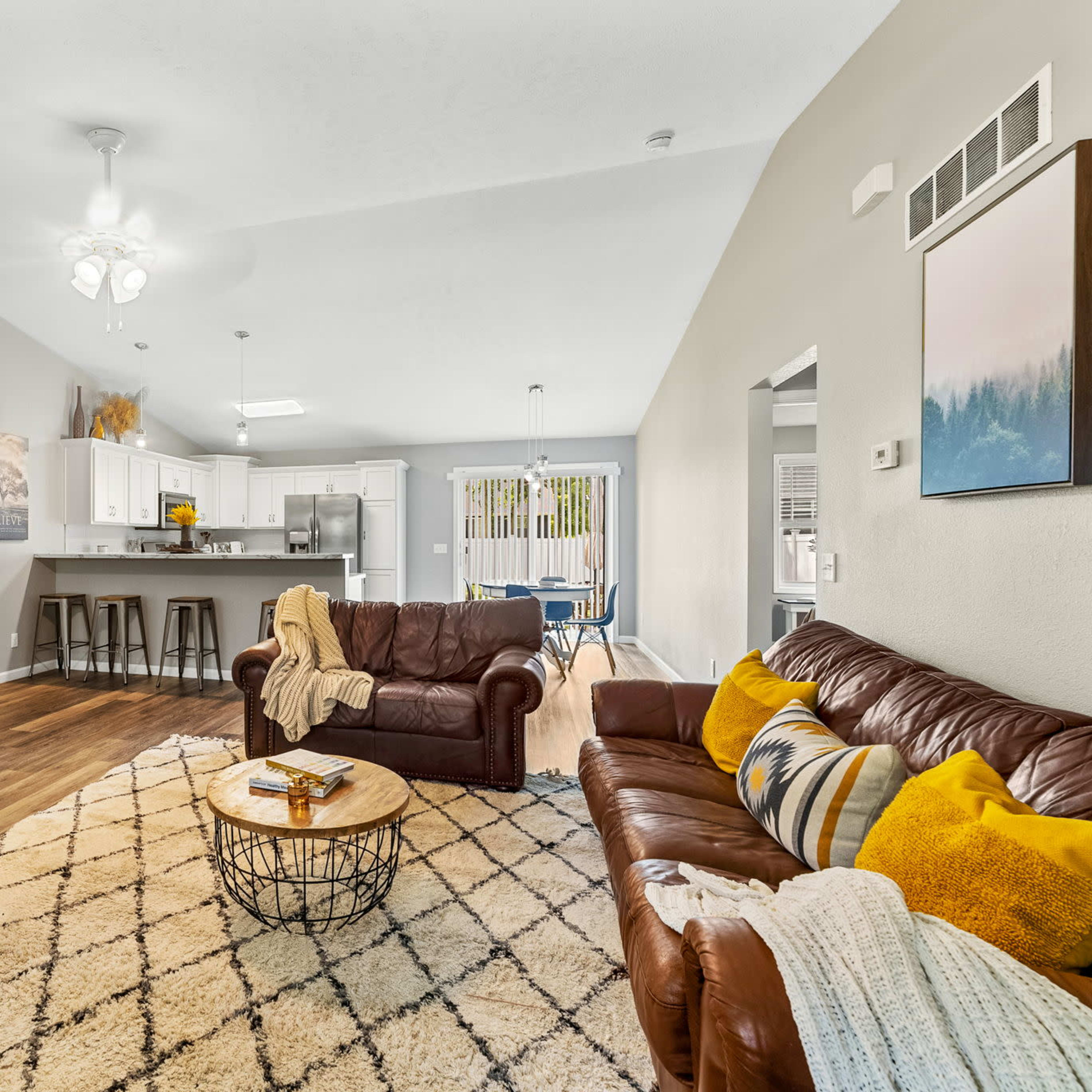 The living room features a brown leather sofa and an adjacent round coffee table, with a fireplace and an open layout leading to a modern kitchen.