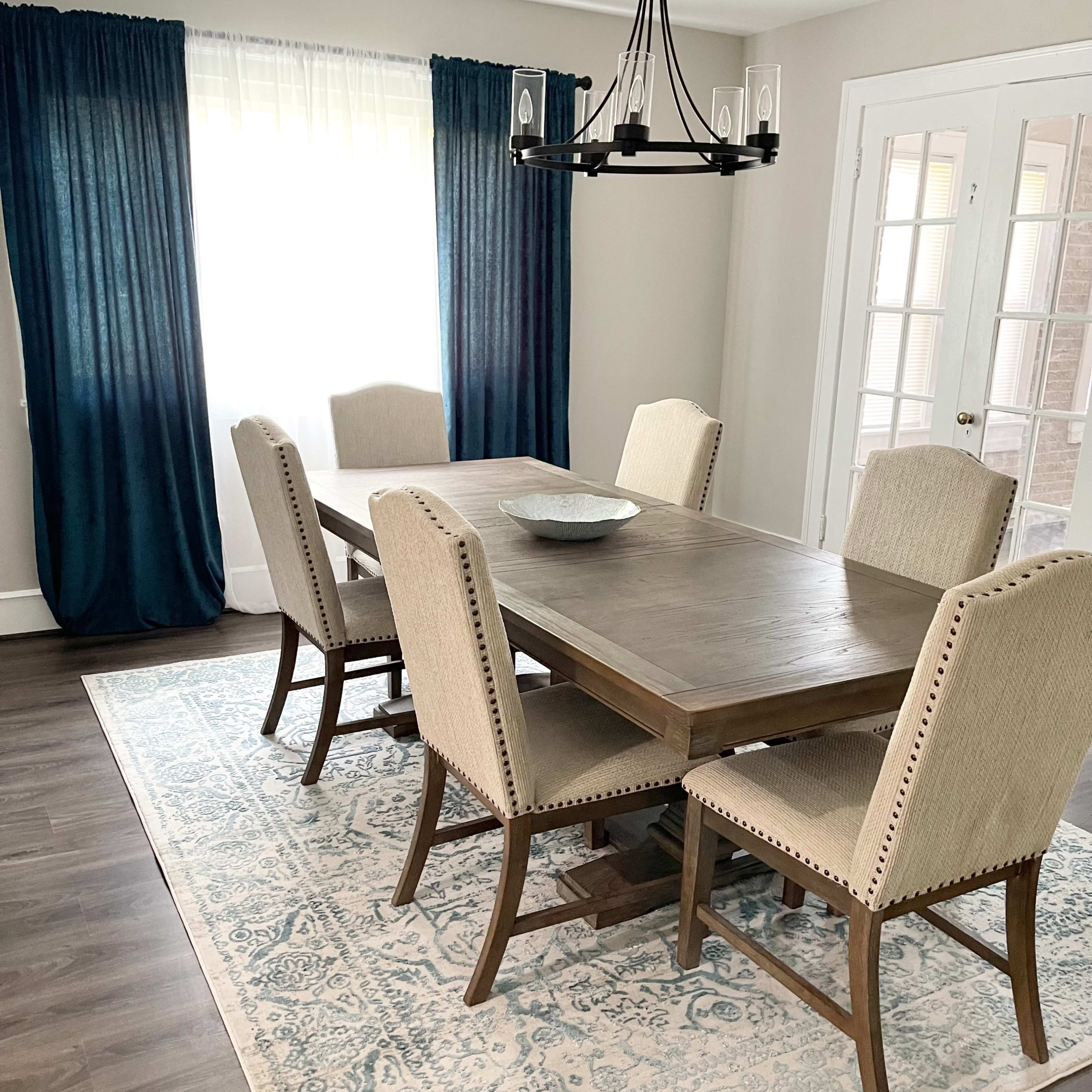 A dining room features a wooden table with six upholstered chairs, a large area rug, and a chandelier above, complemented by sheer curtains.