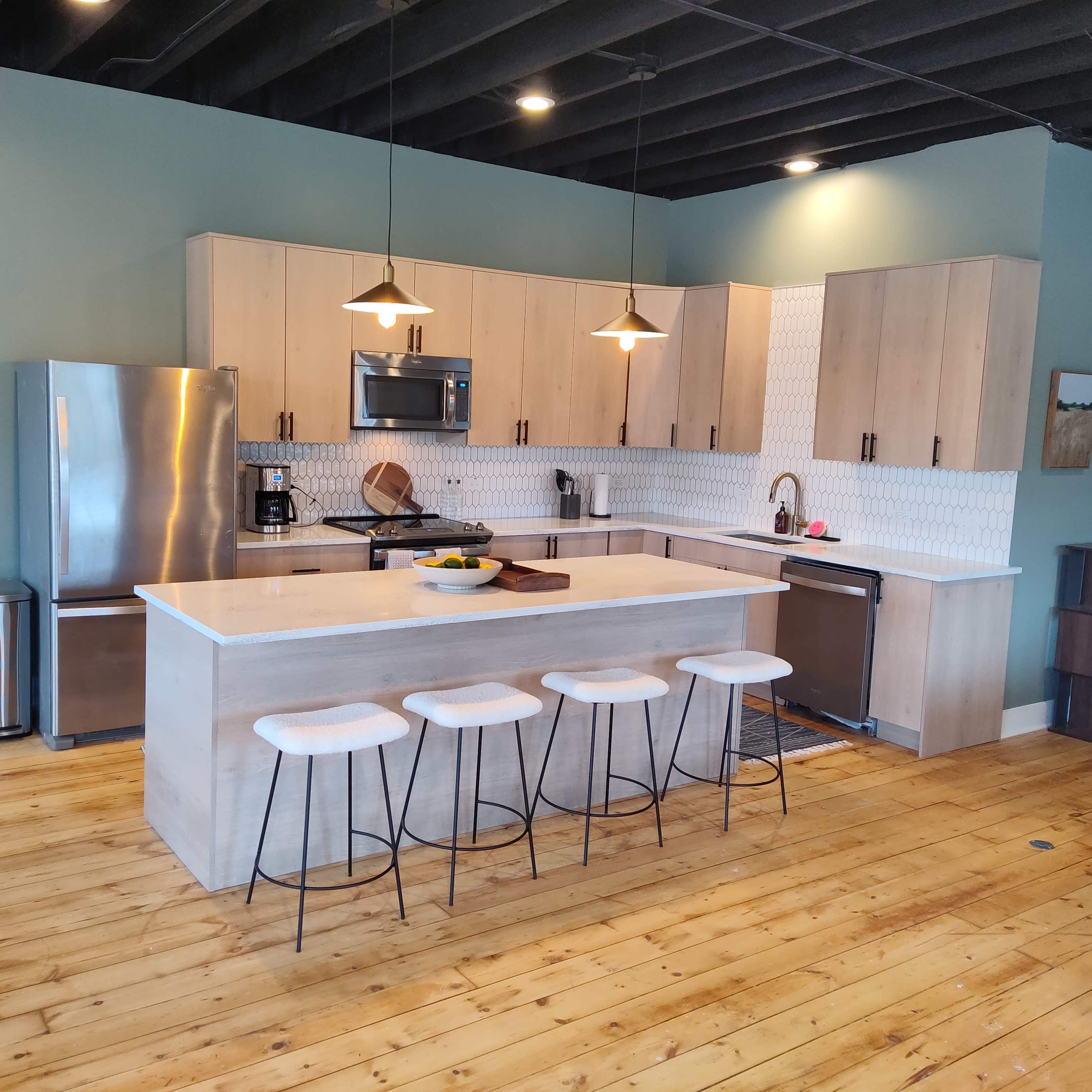 A modern kitchen featuring light wood cabinets, a large island with seating, stainless steel appliances, and a green accent wall.