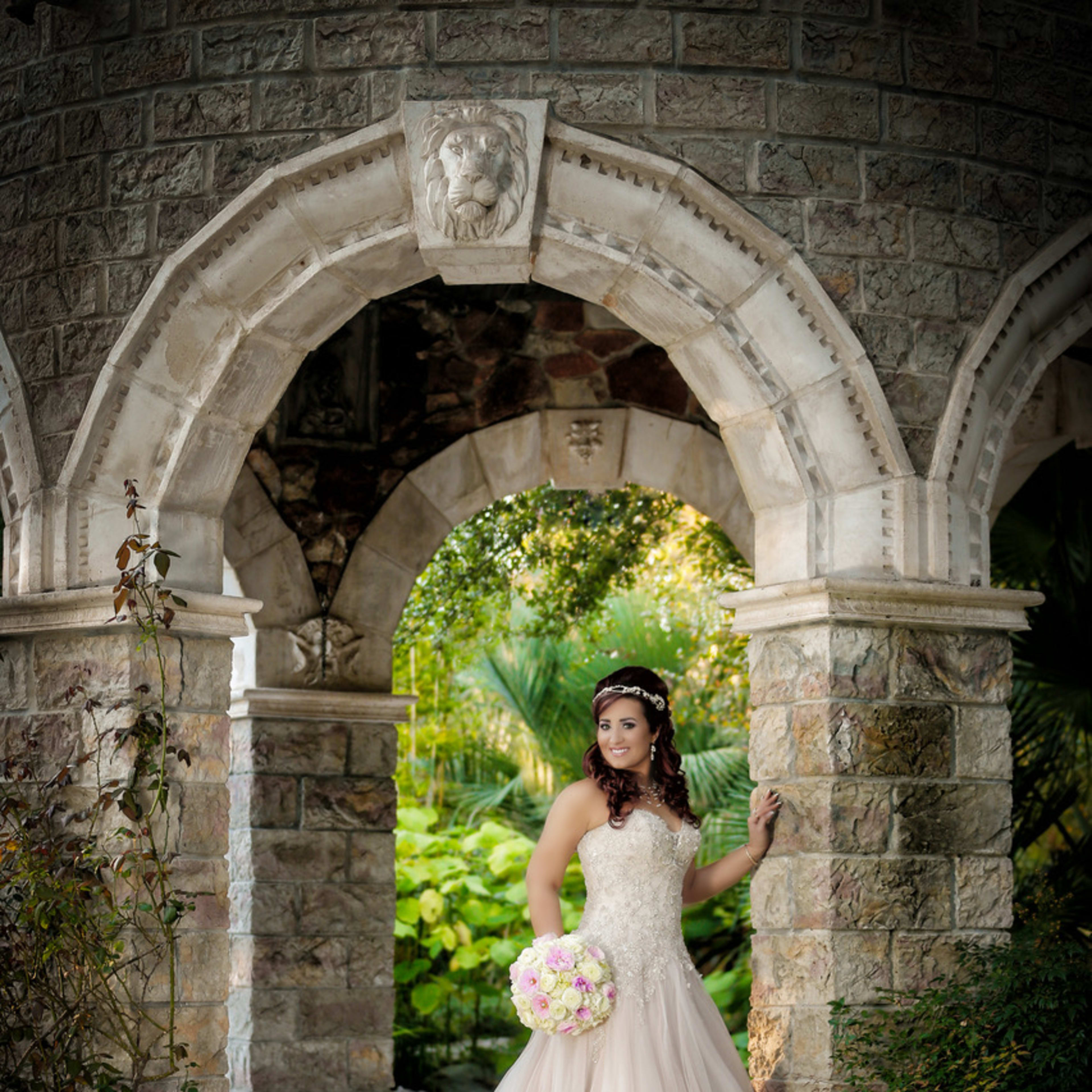 A bride in a flowing gown poses under an ornate stone archway while holding a bouquet of flowers.