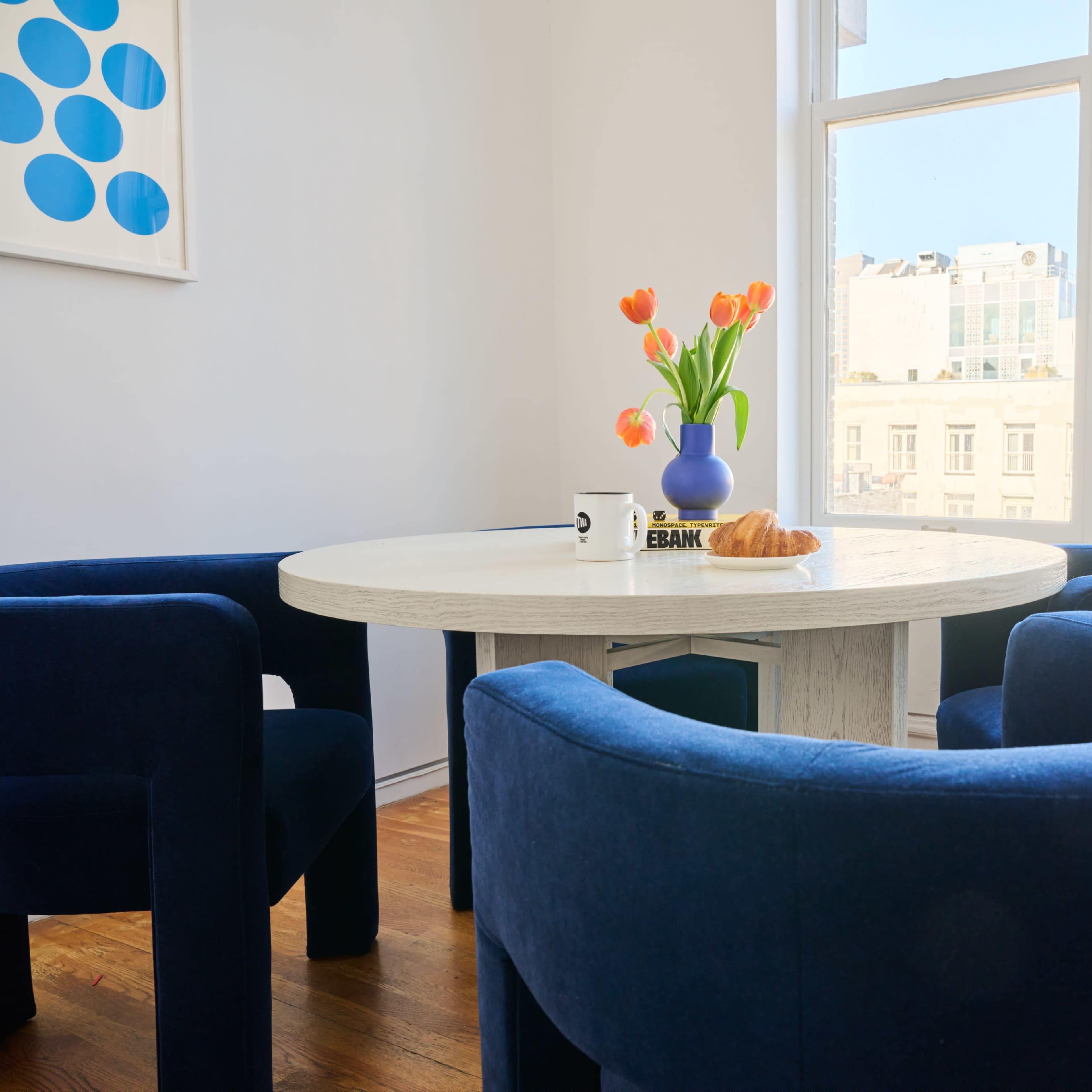 A dining area features a round white table surrounded by four blue velvet chairs, with a vase of tulips and a croissant on the table, and a view of the Empire State Building through the windows.