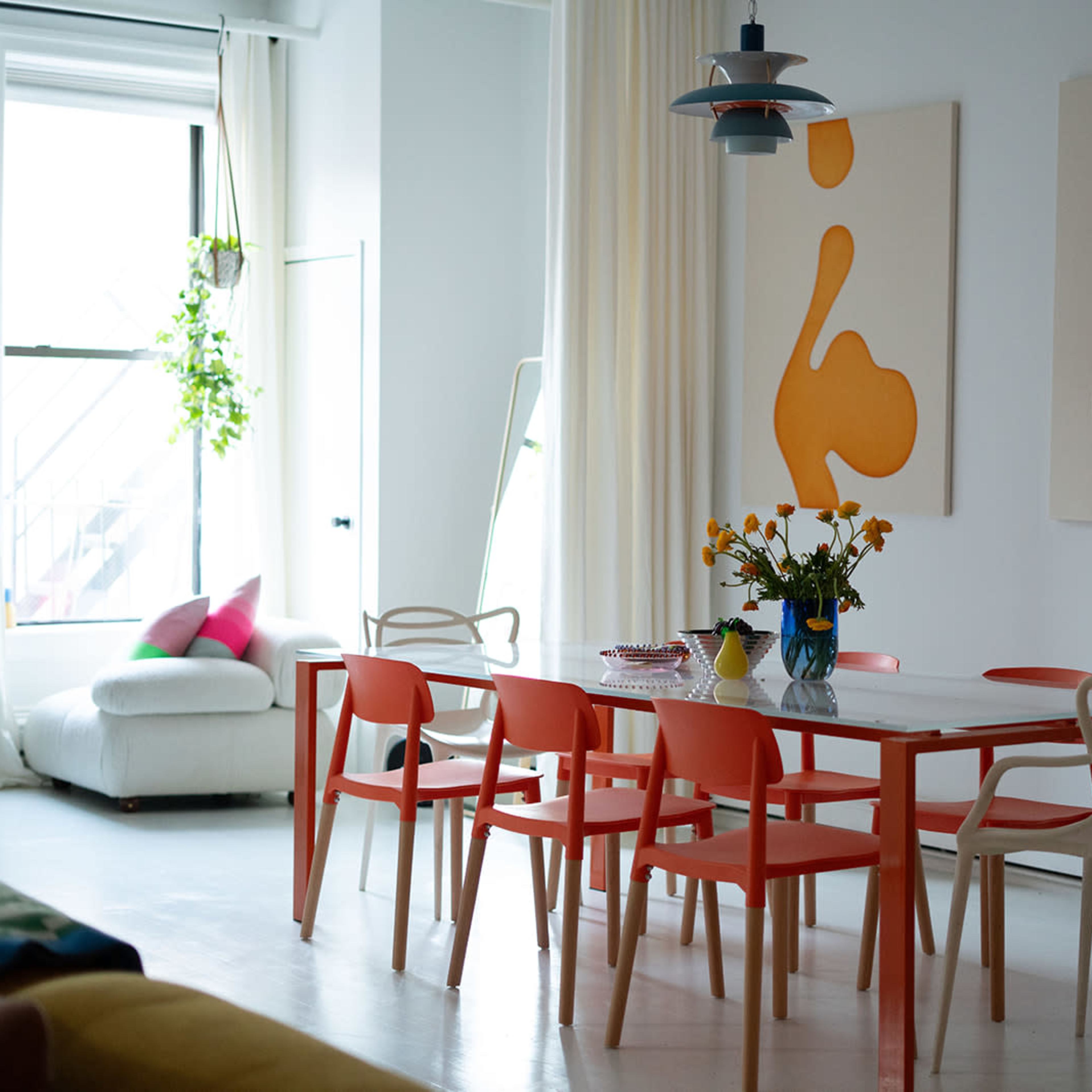 A dining room features a white table surrounded by orange and white chairs, with abstract art on the walls and a vase of yellow flowers in the center.