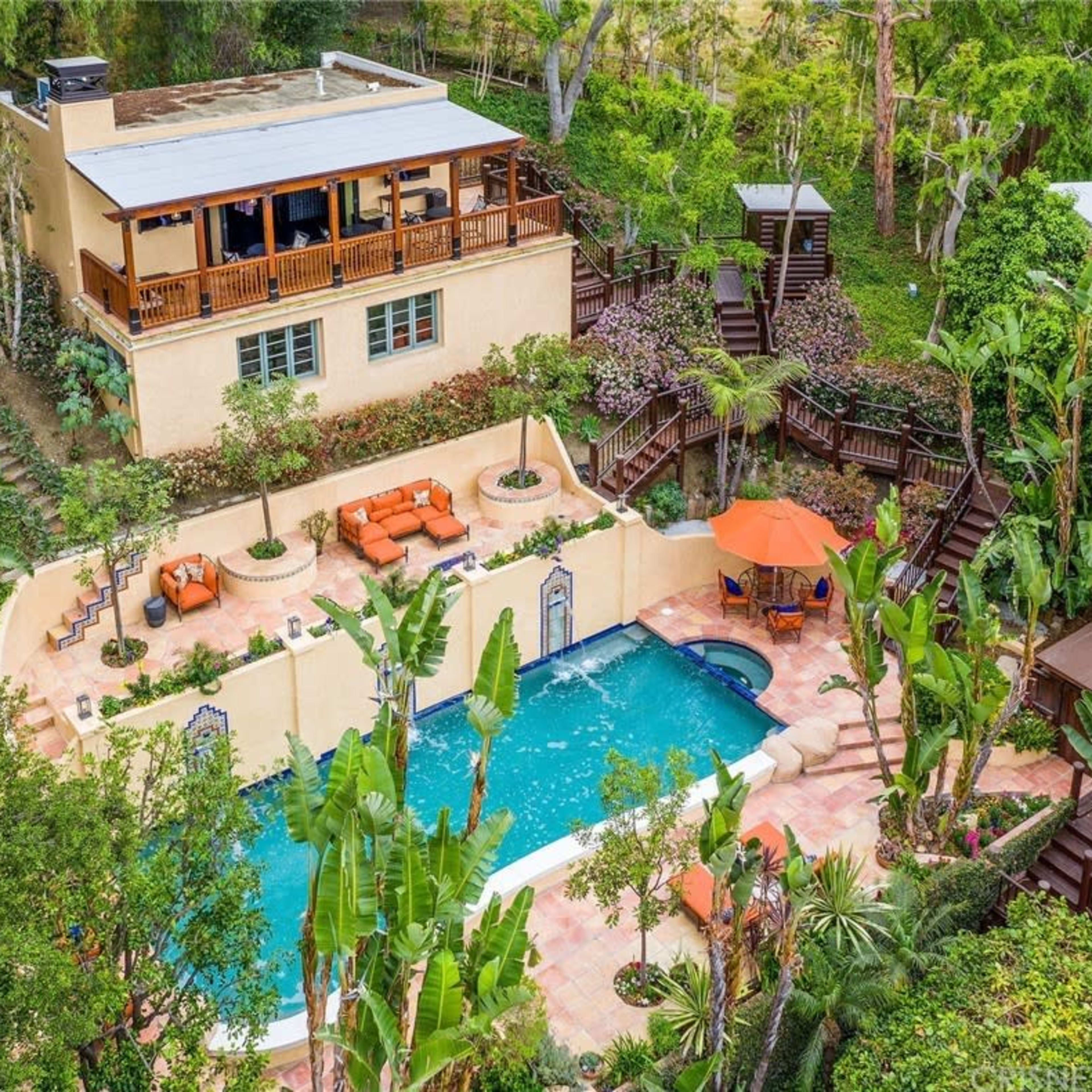 A spacious house with a balcony overlooks a swimming pool surrounded by tropical plants and lounge areas.
