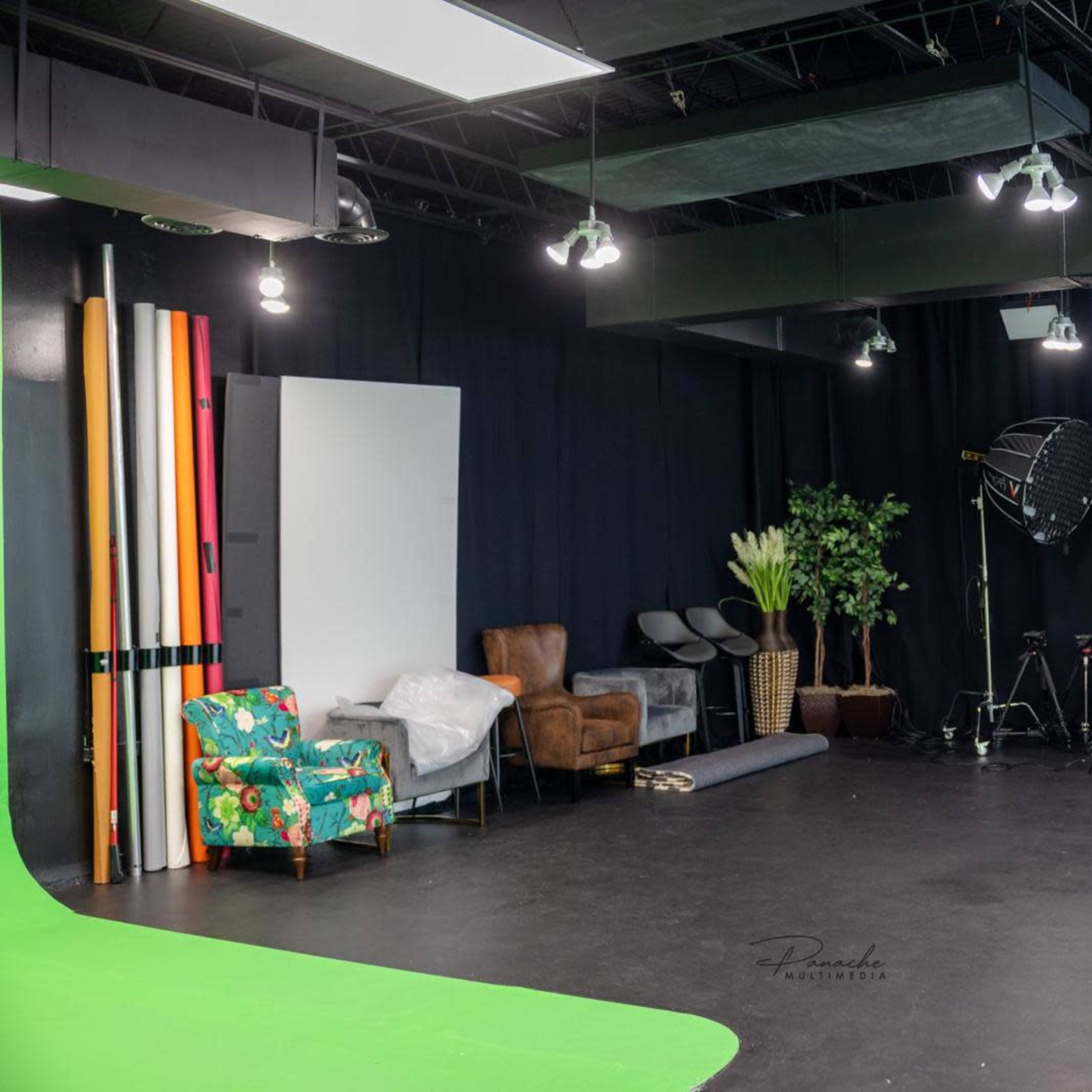 A photography studio features a green backdrop, colorful standing props, lighting equipment, and a mix of furniture including a floral chair and a brown leather sofa.