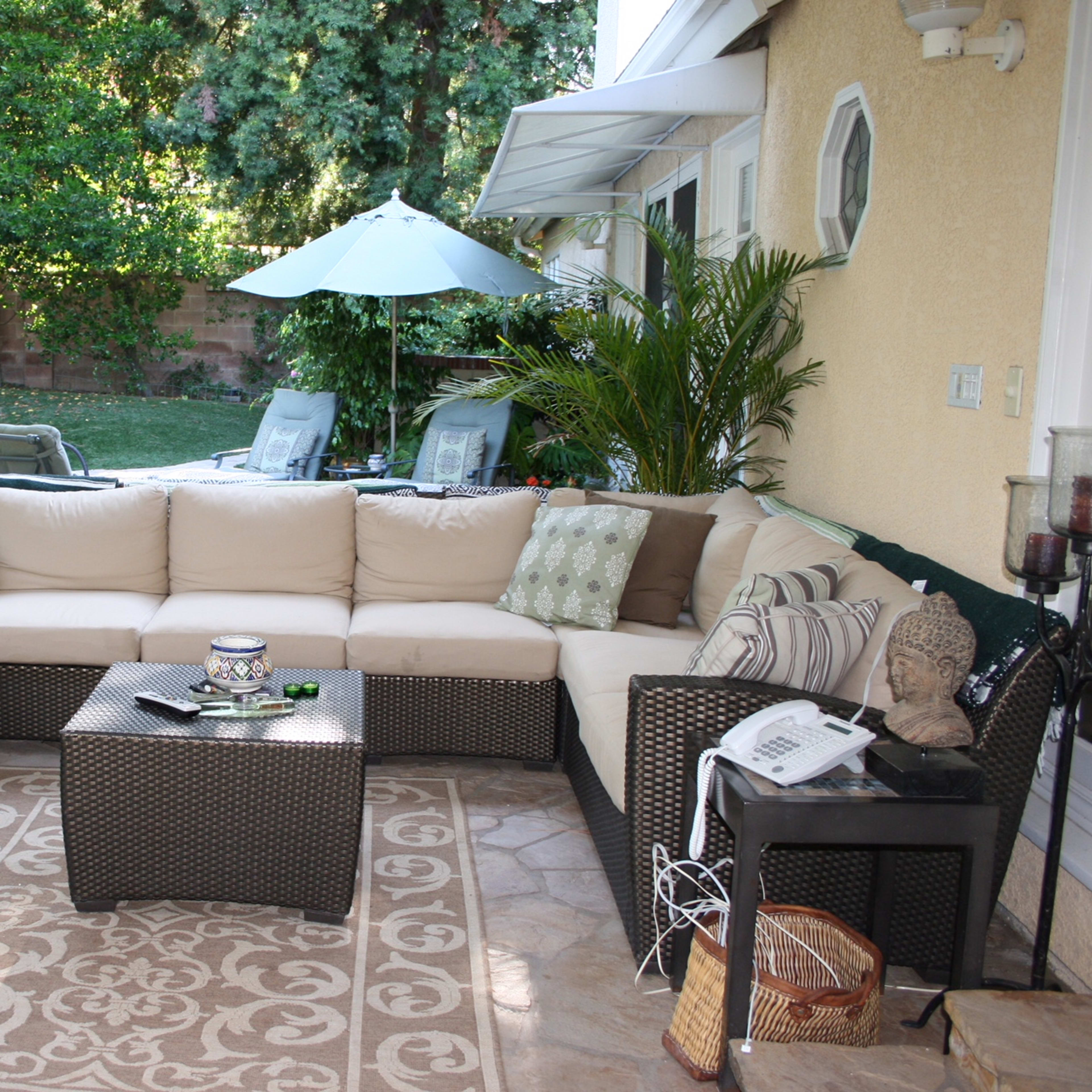 A covered outdoor seating area with a sectional sofa, a coffee table, and a backdrop of a swimming pool and green foliage.