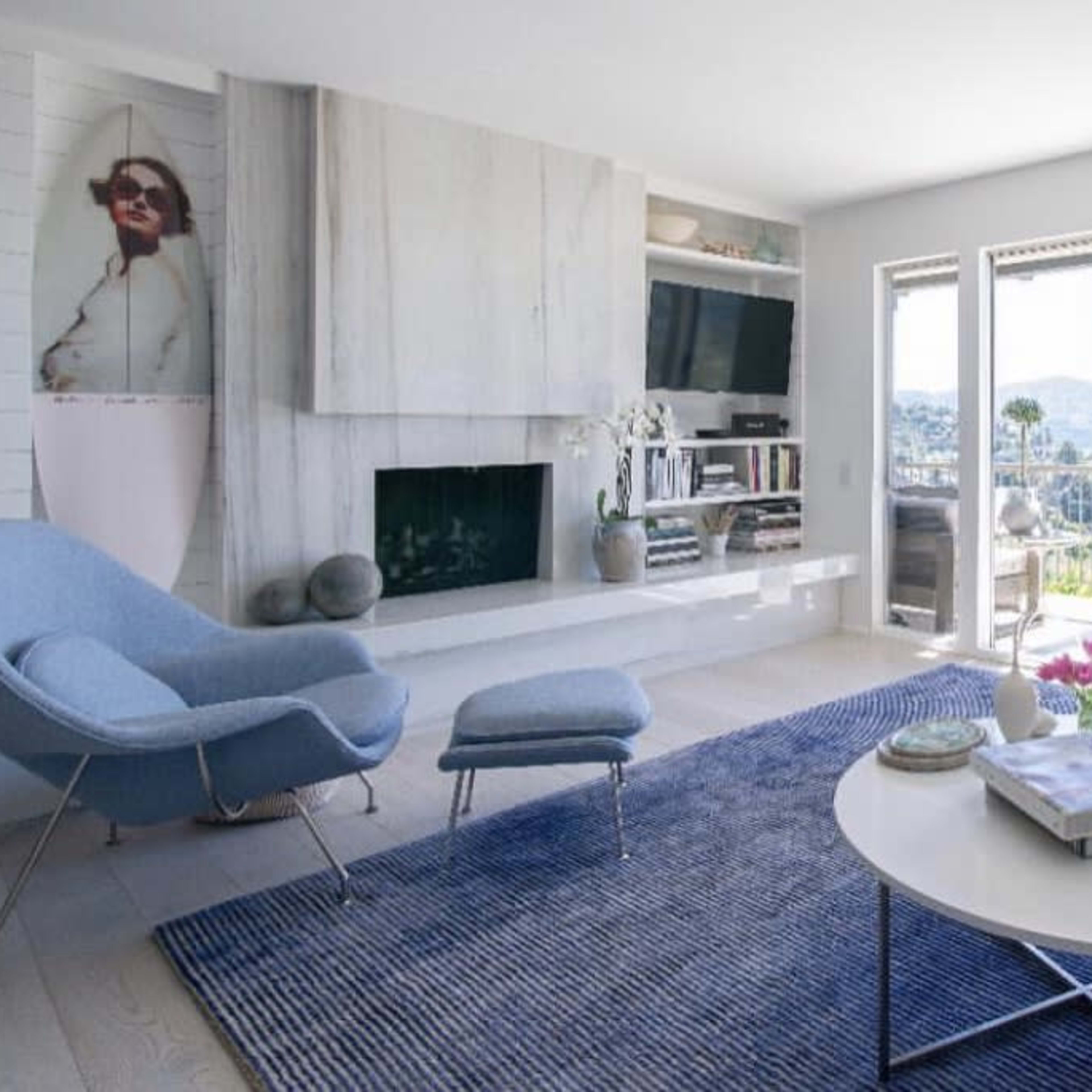 A modern living room with a large window overlooking a landscape, featuring a blue rug, a circular coffee table, a gray chair, and built-in shelves with various decorative items.
