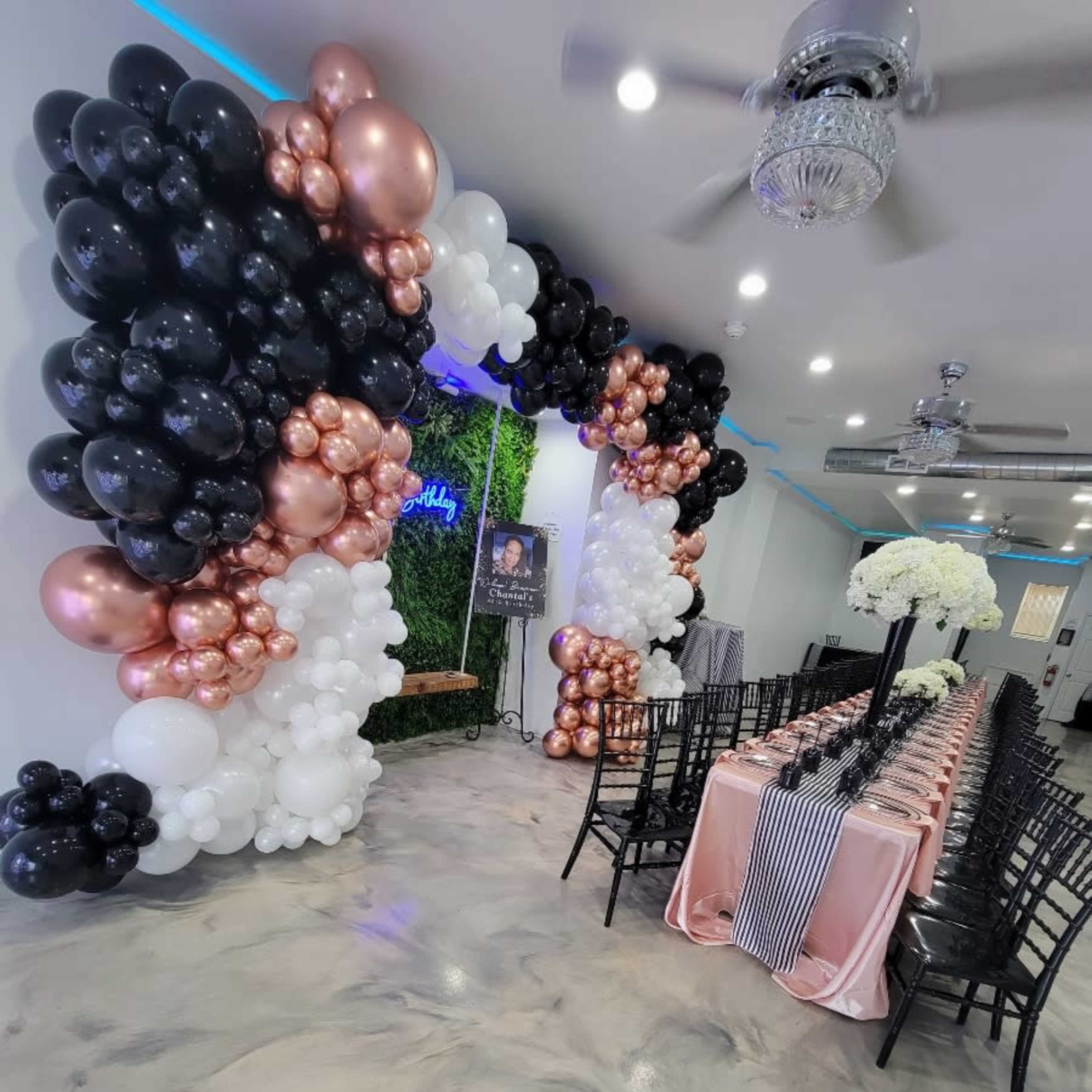 A decorated event space featuring a table set for a celebration, surrounded by balloon arrangements in black, white, and rose gold.