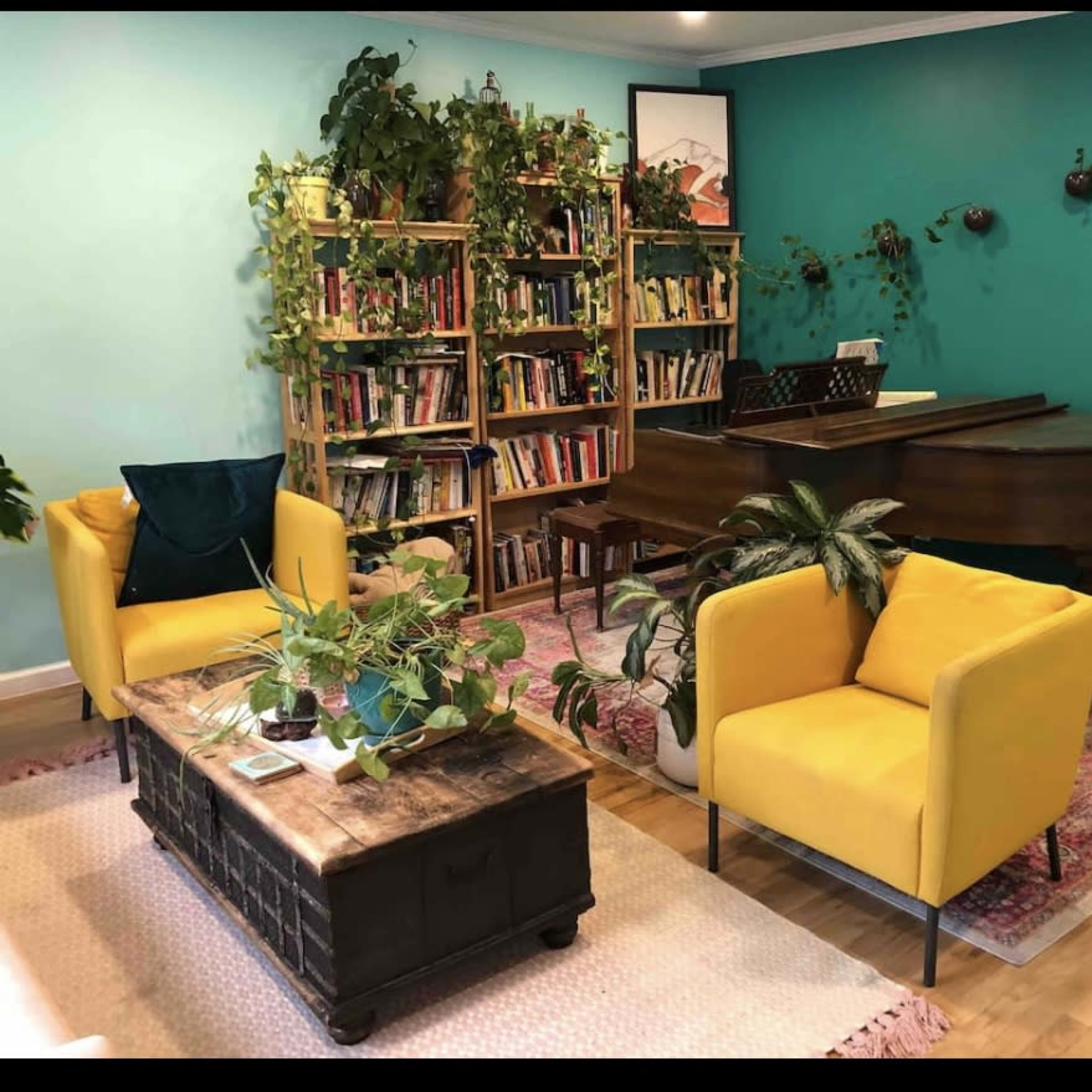 A bright living room features a piano, a bookshelf filled with books, two yellow chairs, and various potted plants against a teal wall.