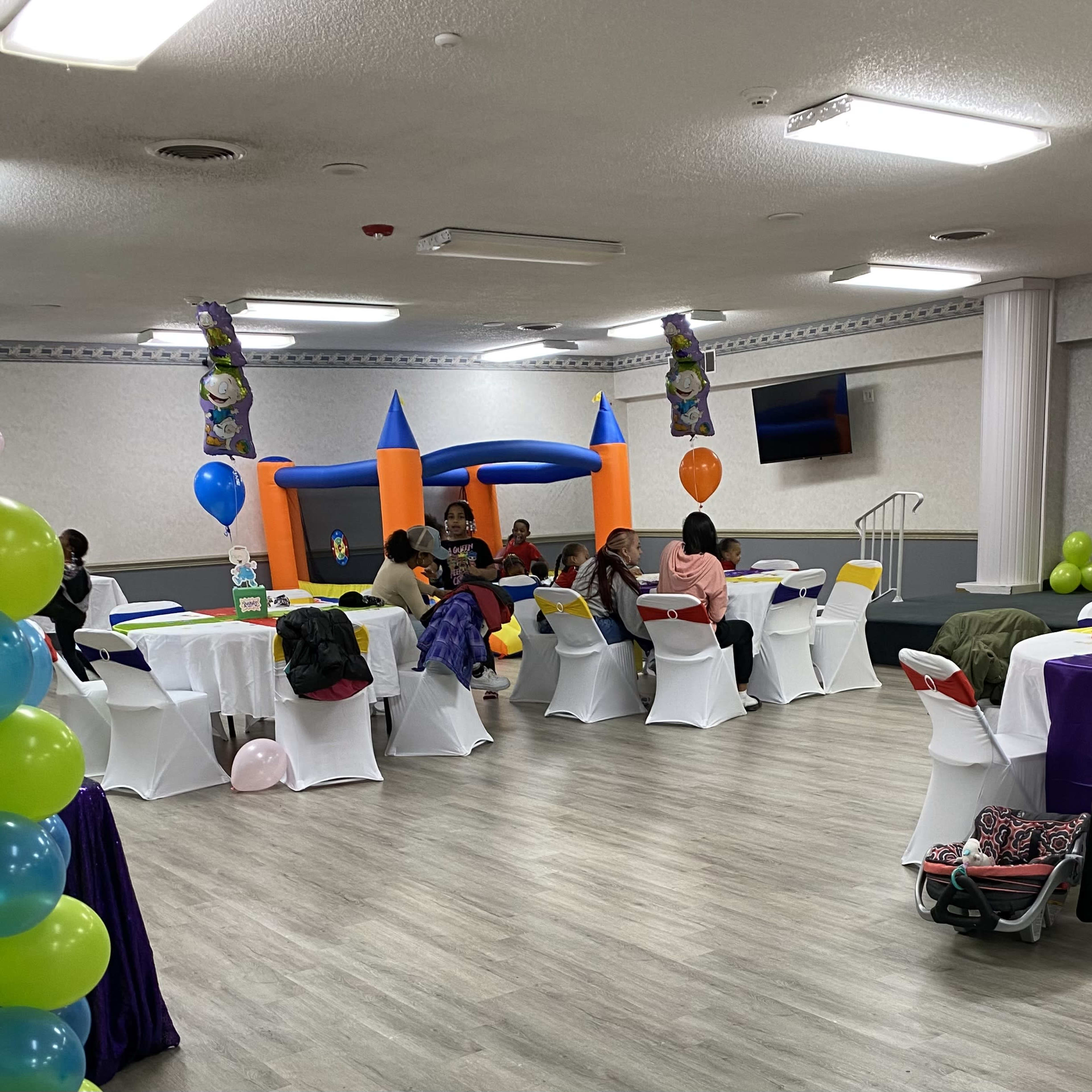 A party or event space with several round tables covered in white tablecloths, colorful decorations, a bounce house in the background, and guests mingling throughout the room.