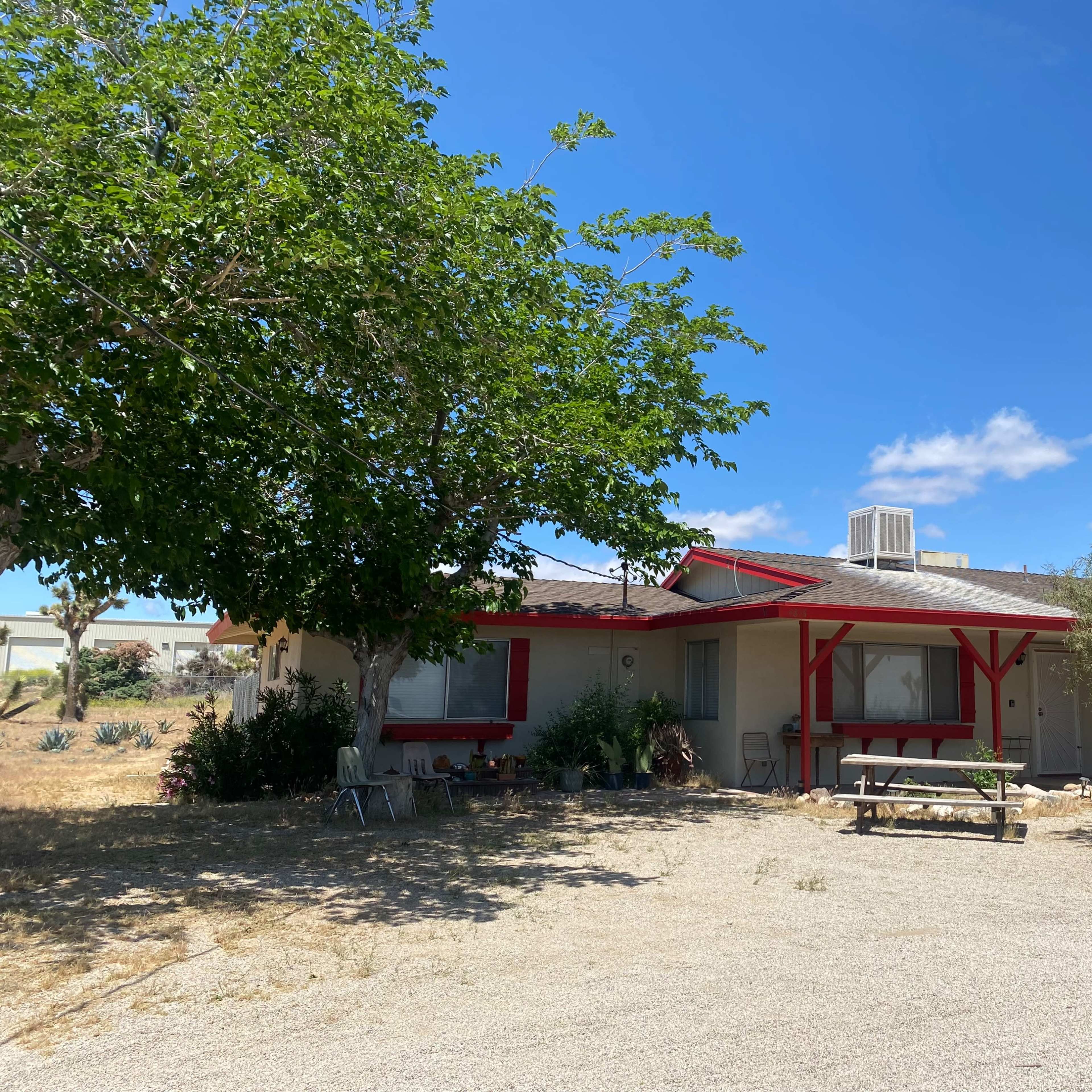 A single-story house with red trim is surrounded by gravel and a few trees in a semi-arid landscape.