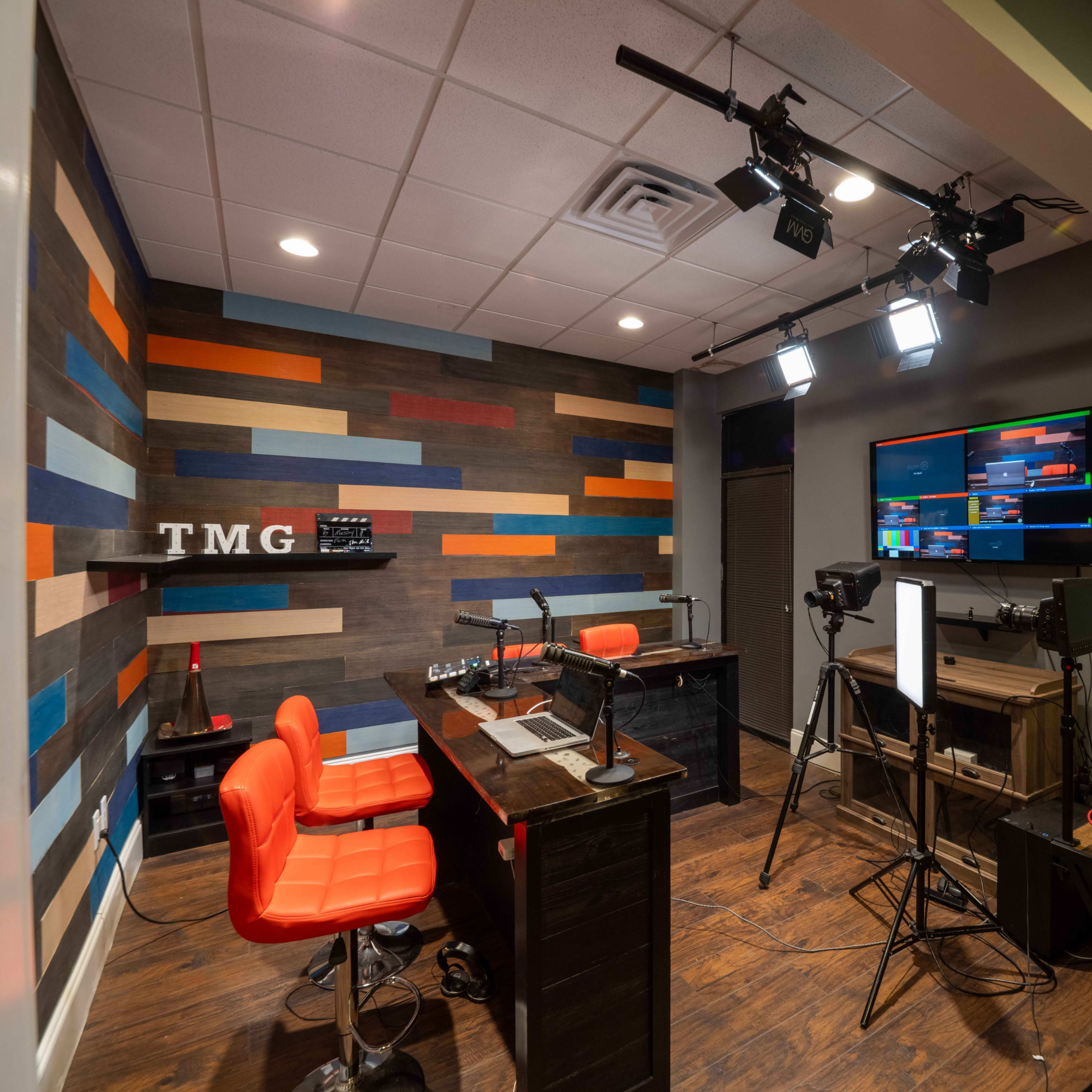 A studio setup with a wooden wall featuring colorful stripes, two orange chairs at a desk, and equipment for audio and video broadcasting.