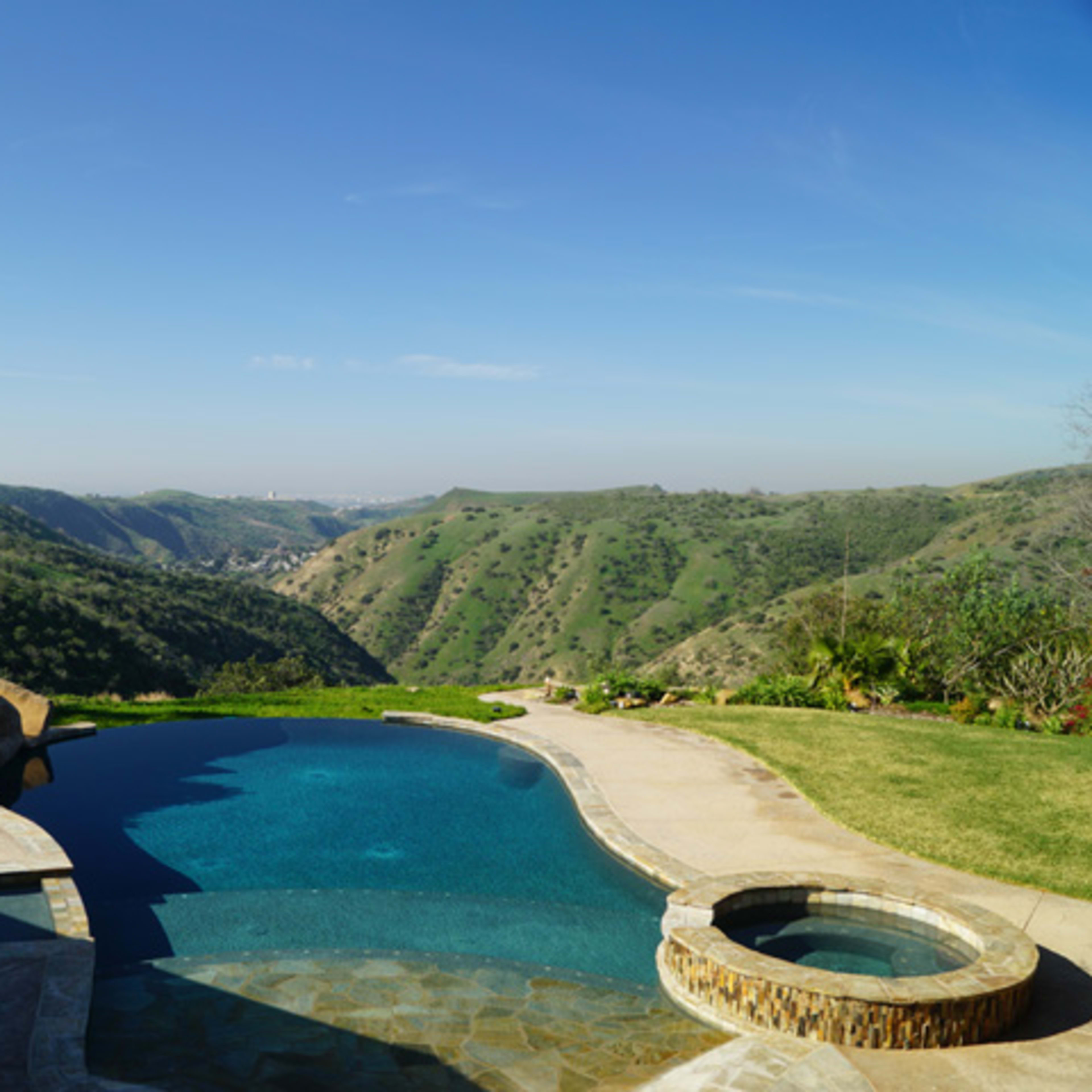 A swimming pool with a spa overlooks rolling green hills under a clear blue sky.