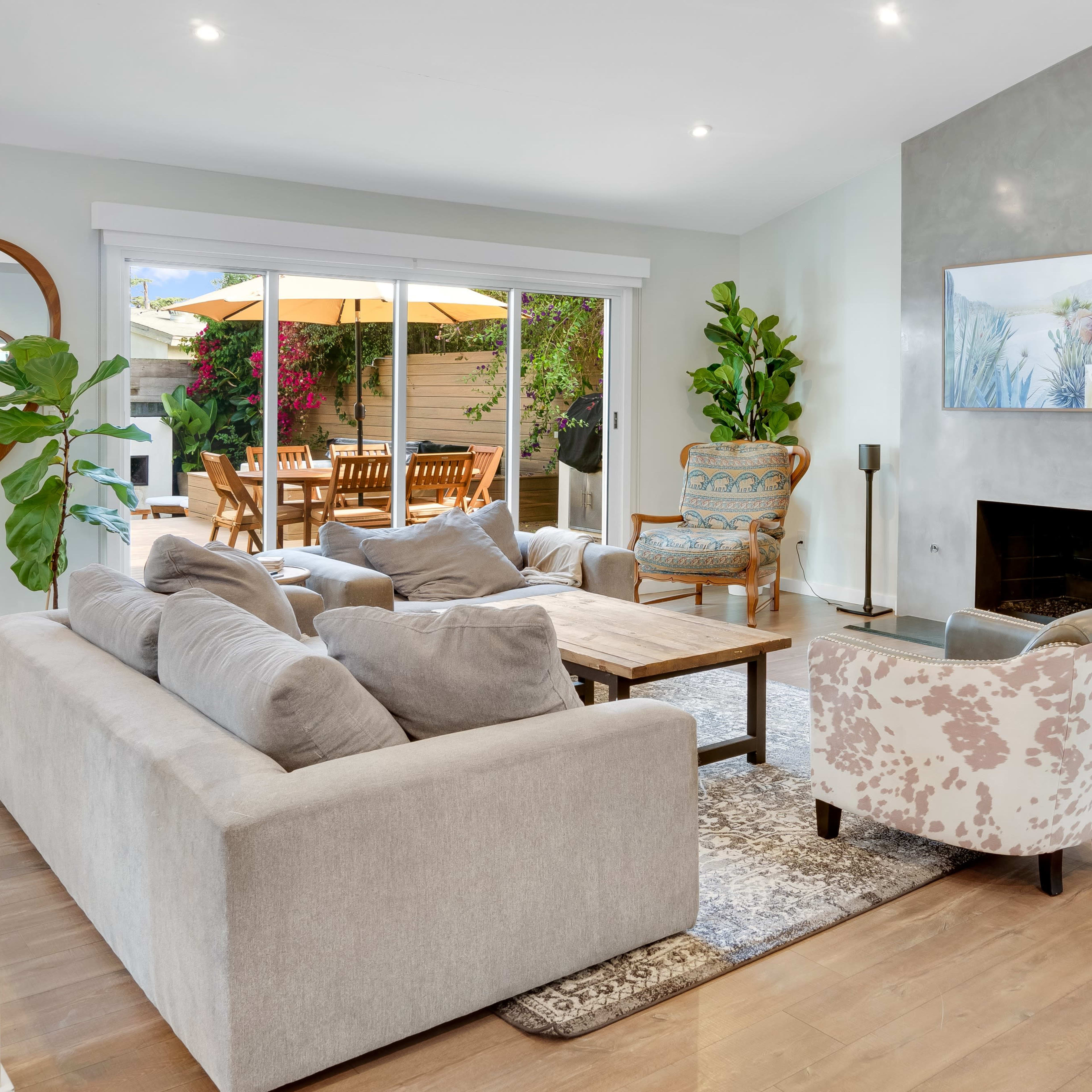 A modern living room features a light gray sofa, a wooden coffee table, and a large window overlooking a patio with outdoor seating.