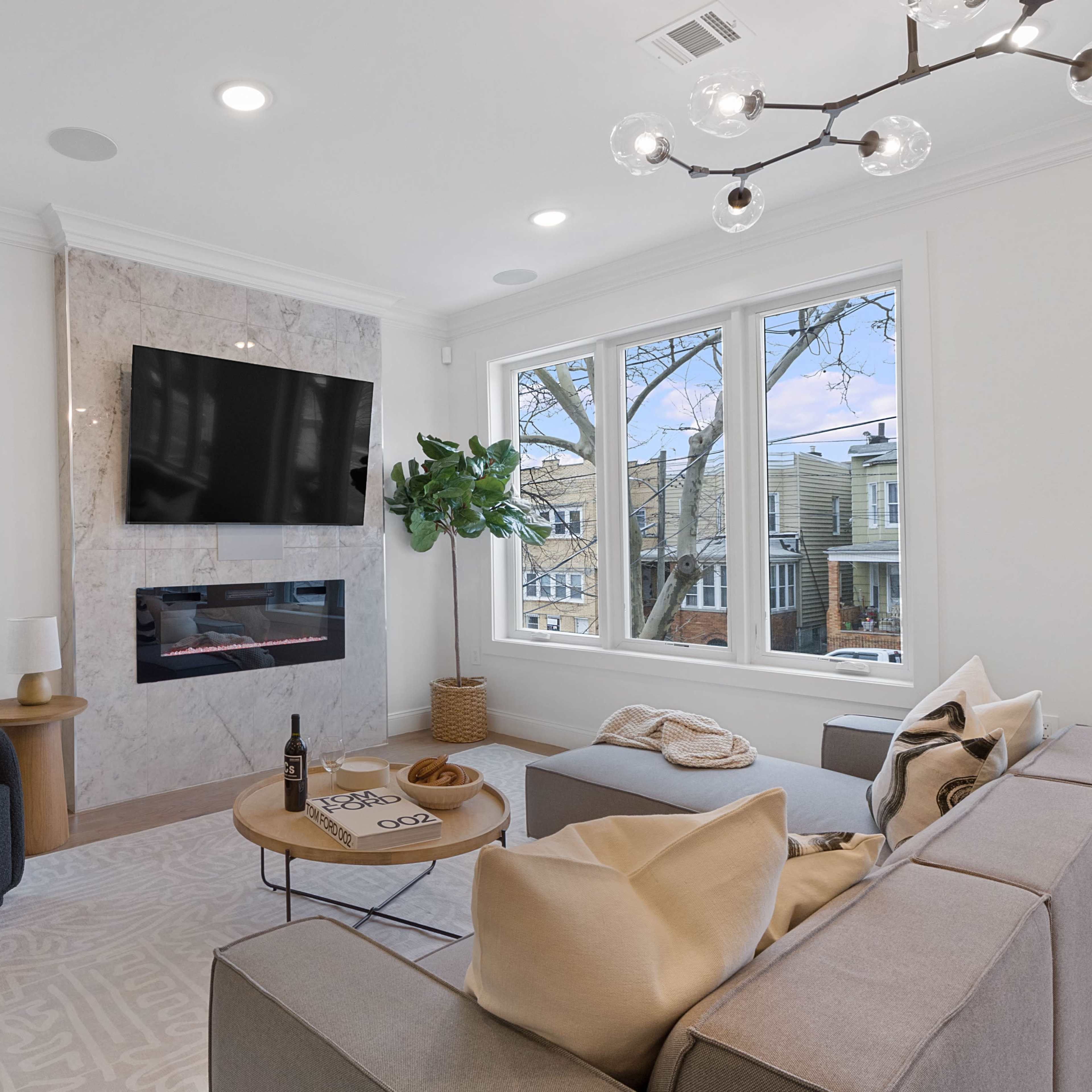 A modern living room features a large wall-mounted television, a minimalist fireplace, a dark chair, a sectional sofa with cushions, and a round coffee table.