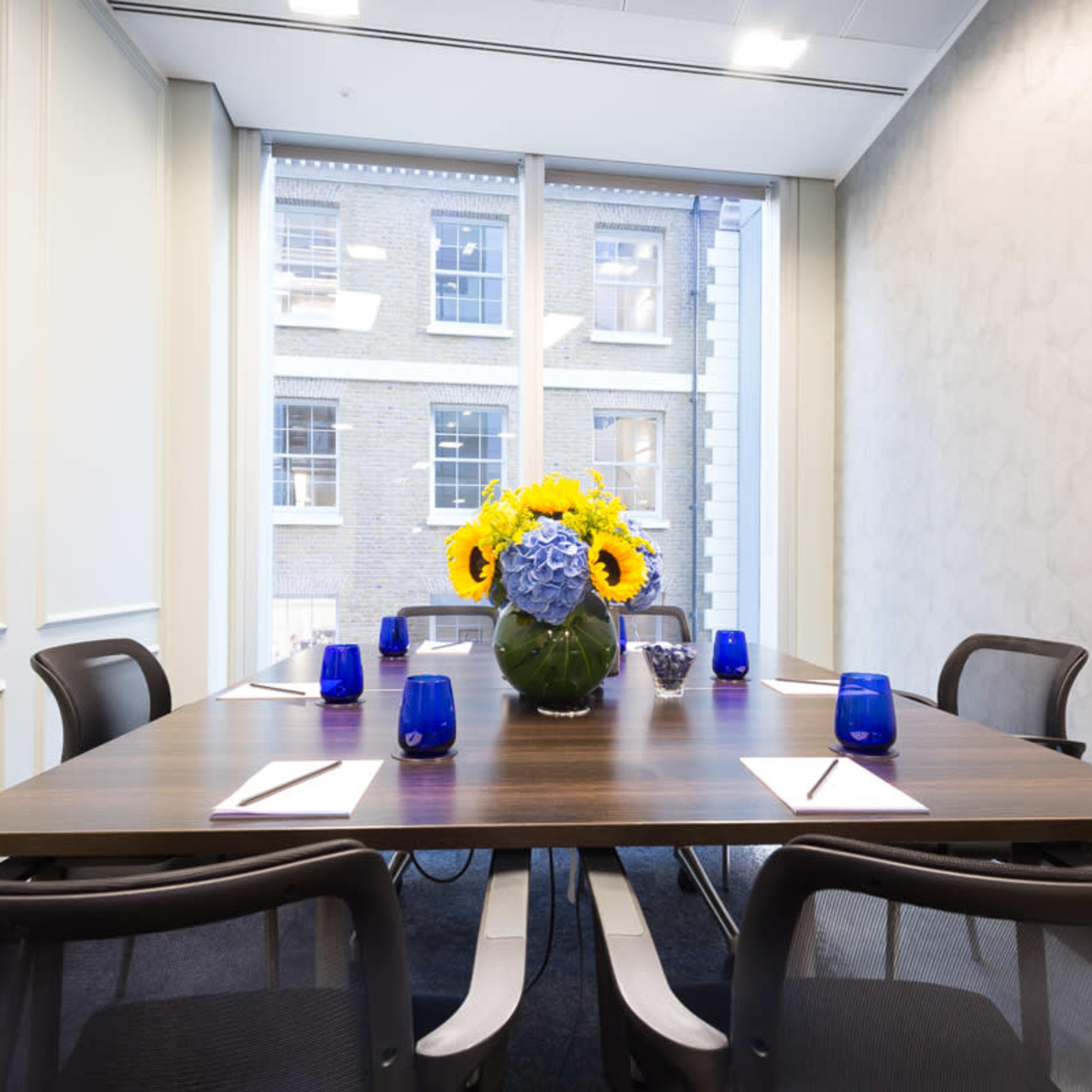 A meeting room features a wooden table set with notepads and blue glasses, centered around a vase of yellow flowers.