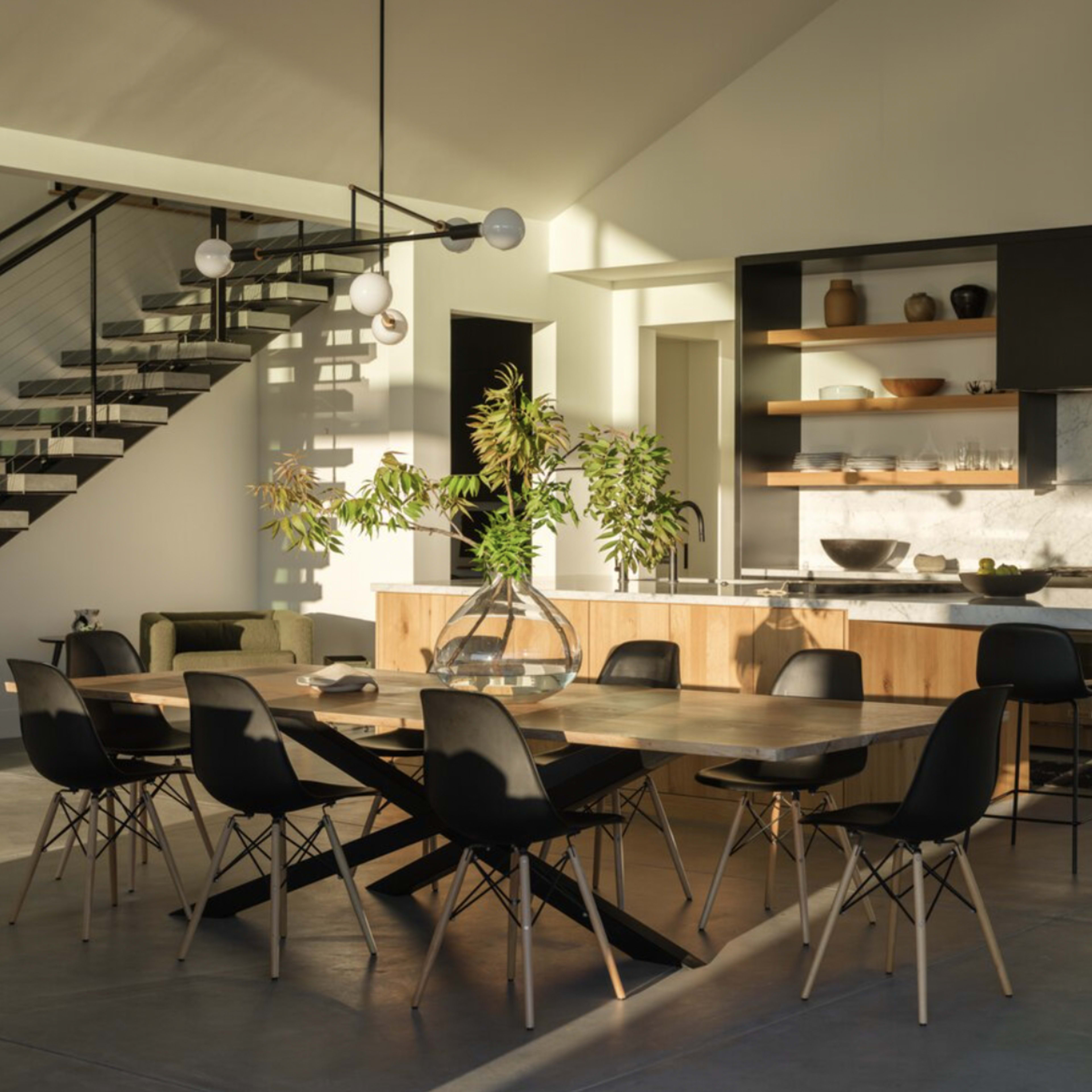 A spacious, modern dining area features a large table surrounded by black chairs, with a staircase and a kitchen area visible in the background.