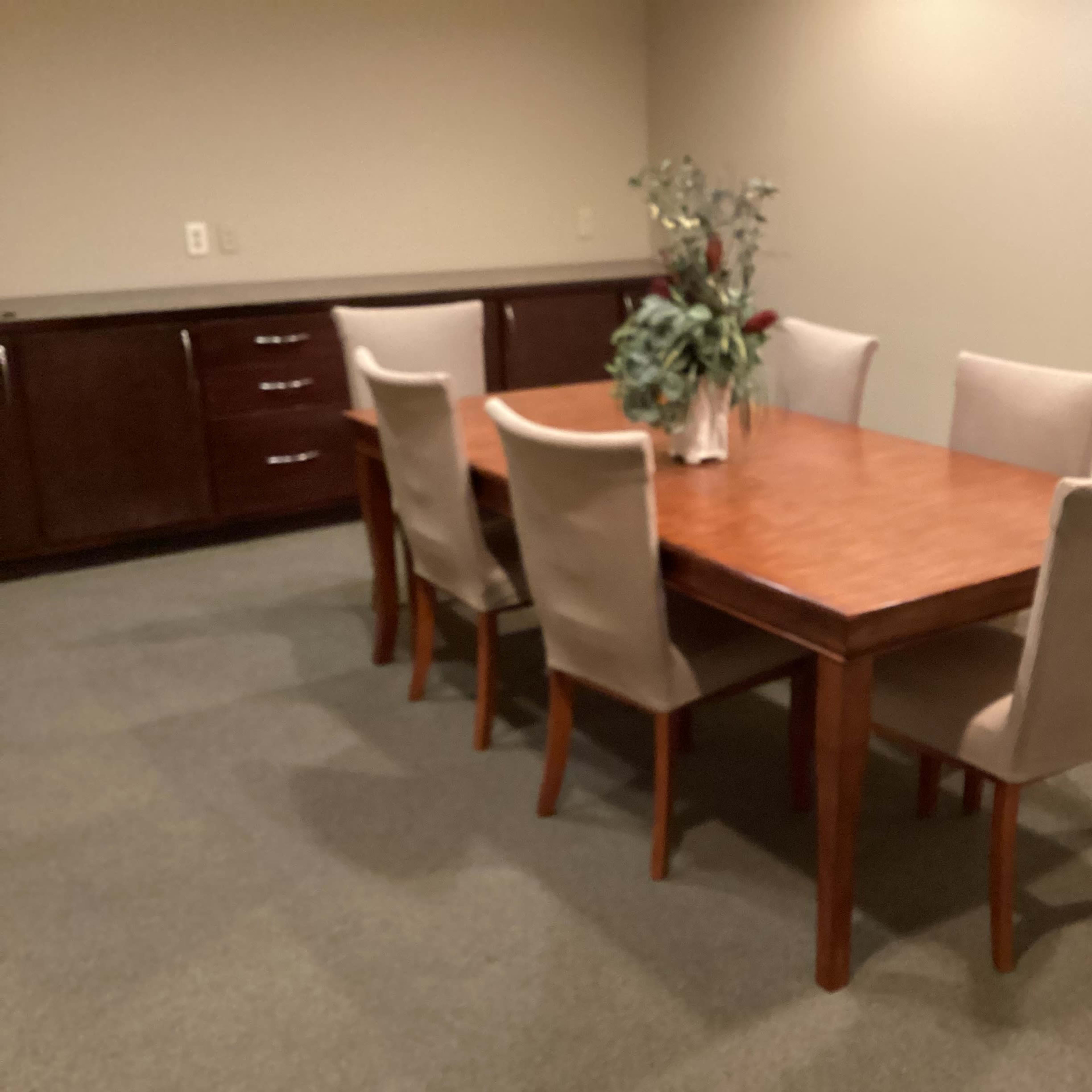 A wooden dining table is set in a room surrounded by six upholstered chairs, with a vase of flowers in the center.