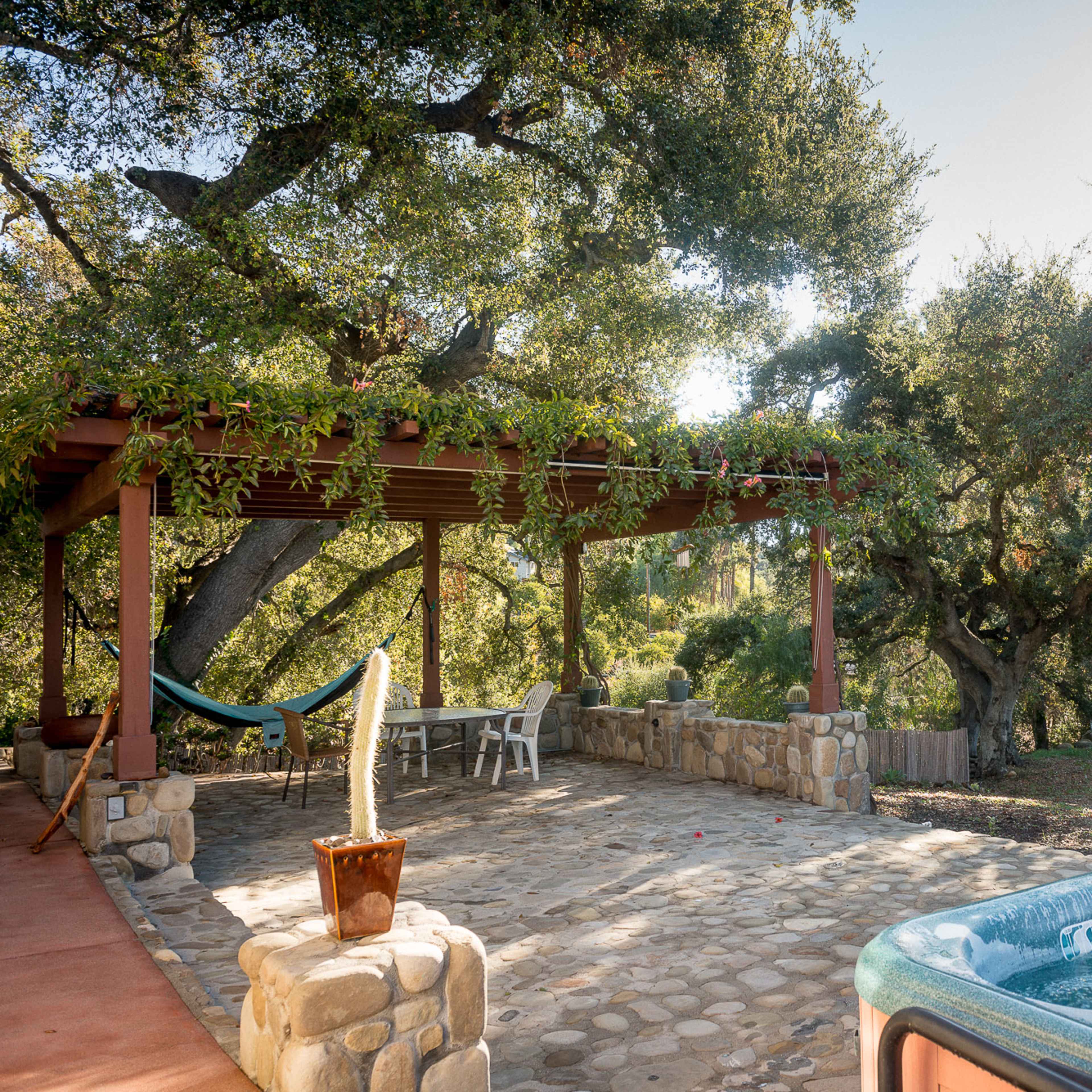 A patio area with a hot tub, a barbecue grill, a stone floor, and a hammock hanging under a wooden pergola surrounded by trees.