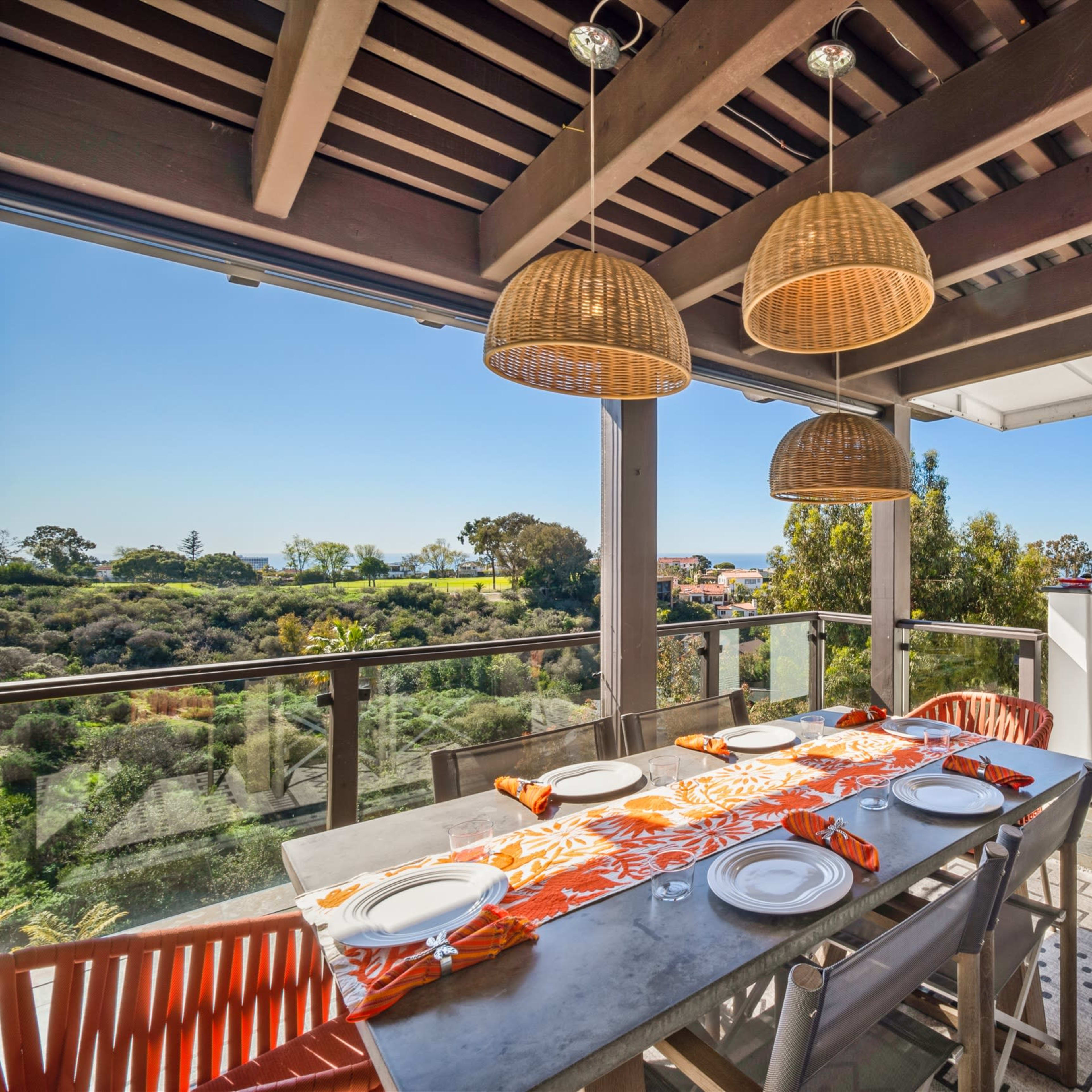 A modern outdoor dining area with a table set for six, overlooking a scenic view of greenery and the ocean.