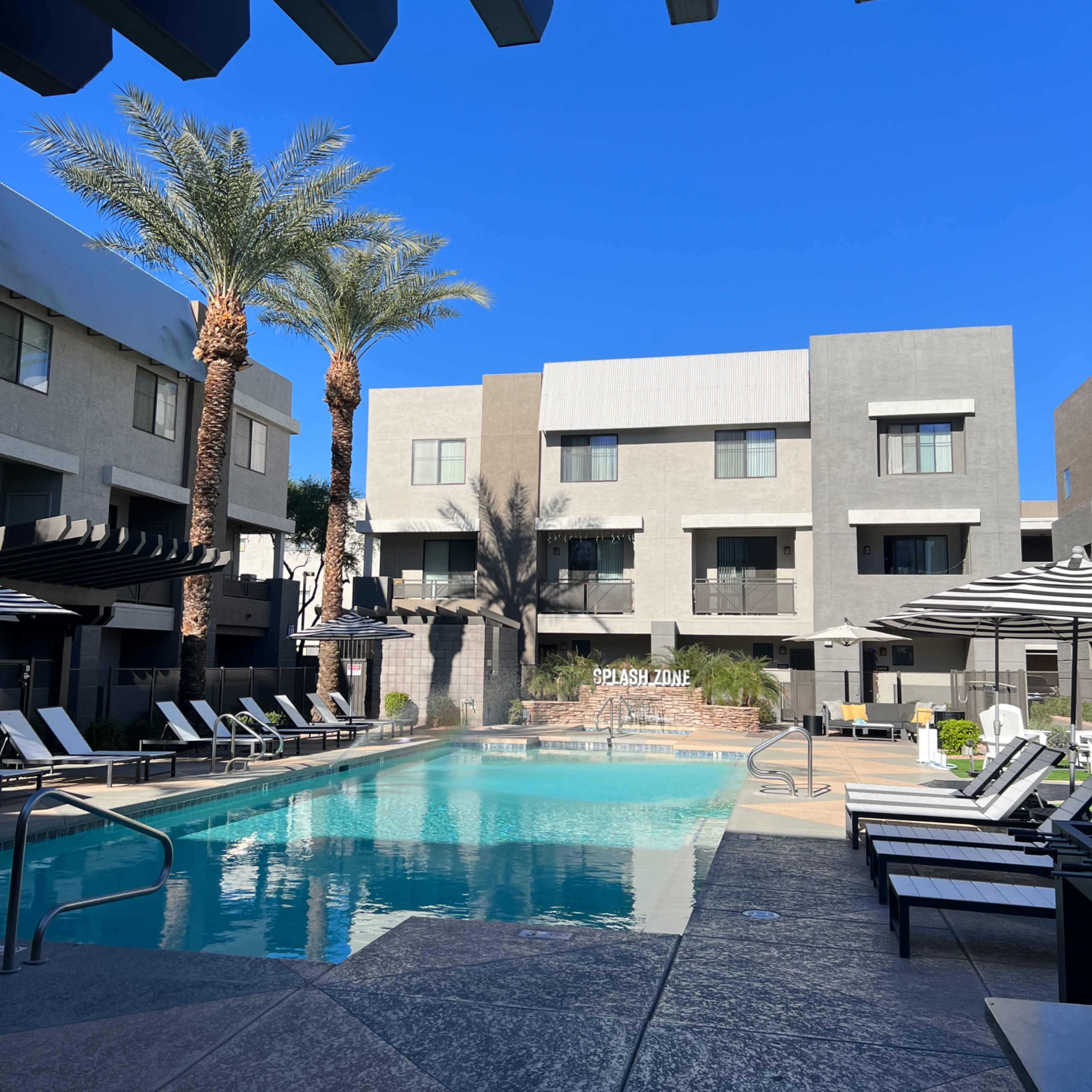 The scene features a swimming pool surrounded by lounge chairs and palm trees, with modern apartment buildings in the background under a clear blue sky.