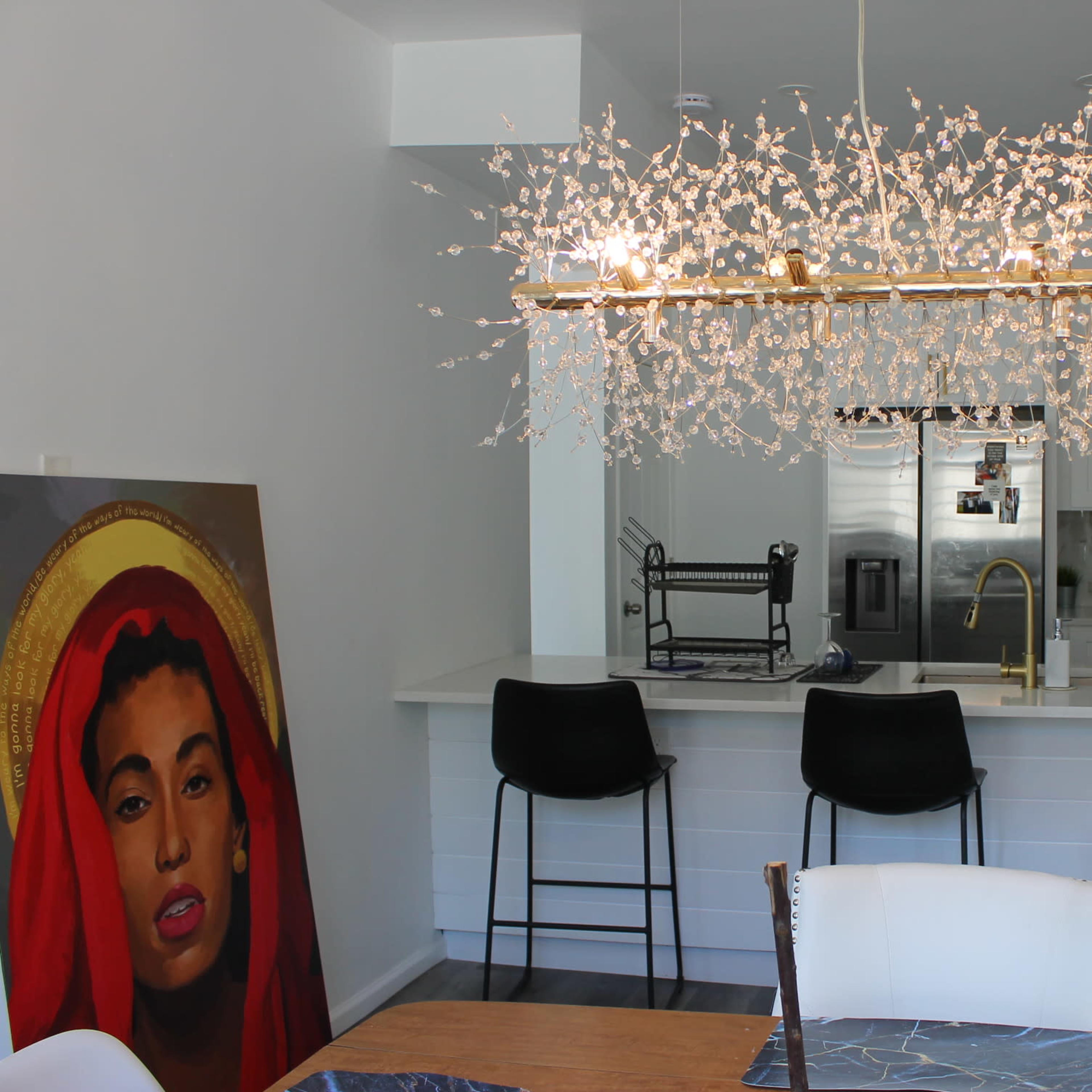 A modern kitchen features a decorative chandelier above a dining table, with a painting of a woman in red on the side.