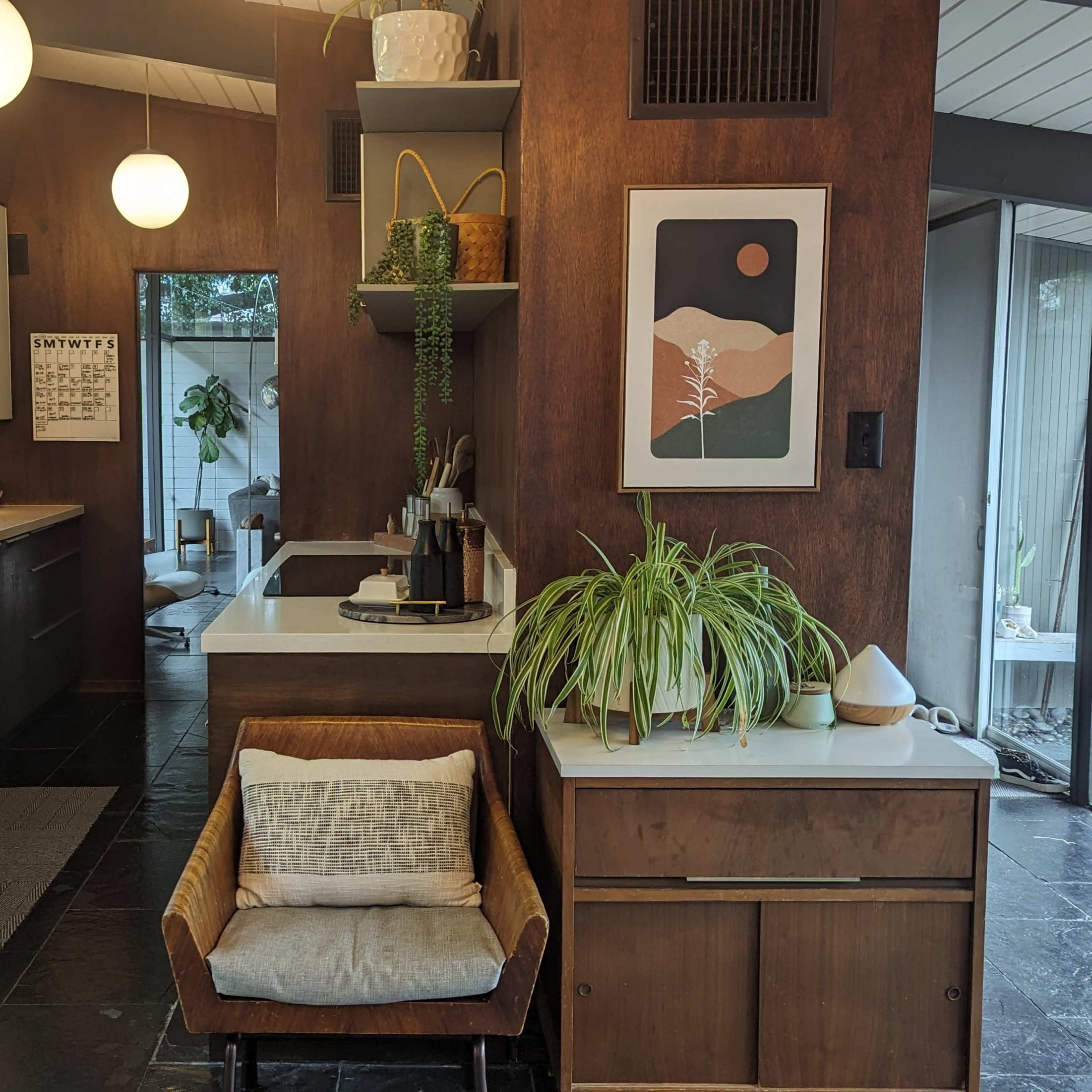 A modern kitchen features wooden cabinetry, a minimalist design, and various plants, with pendant lighting and an art piece on the wall.