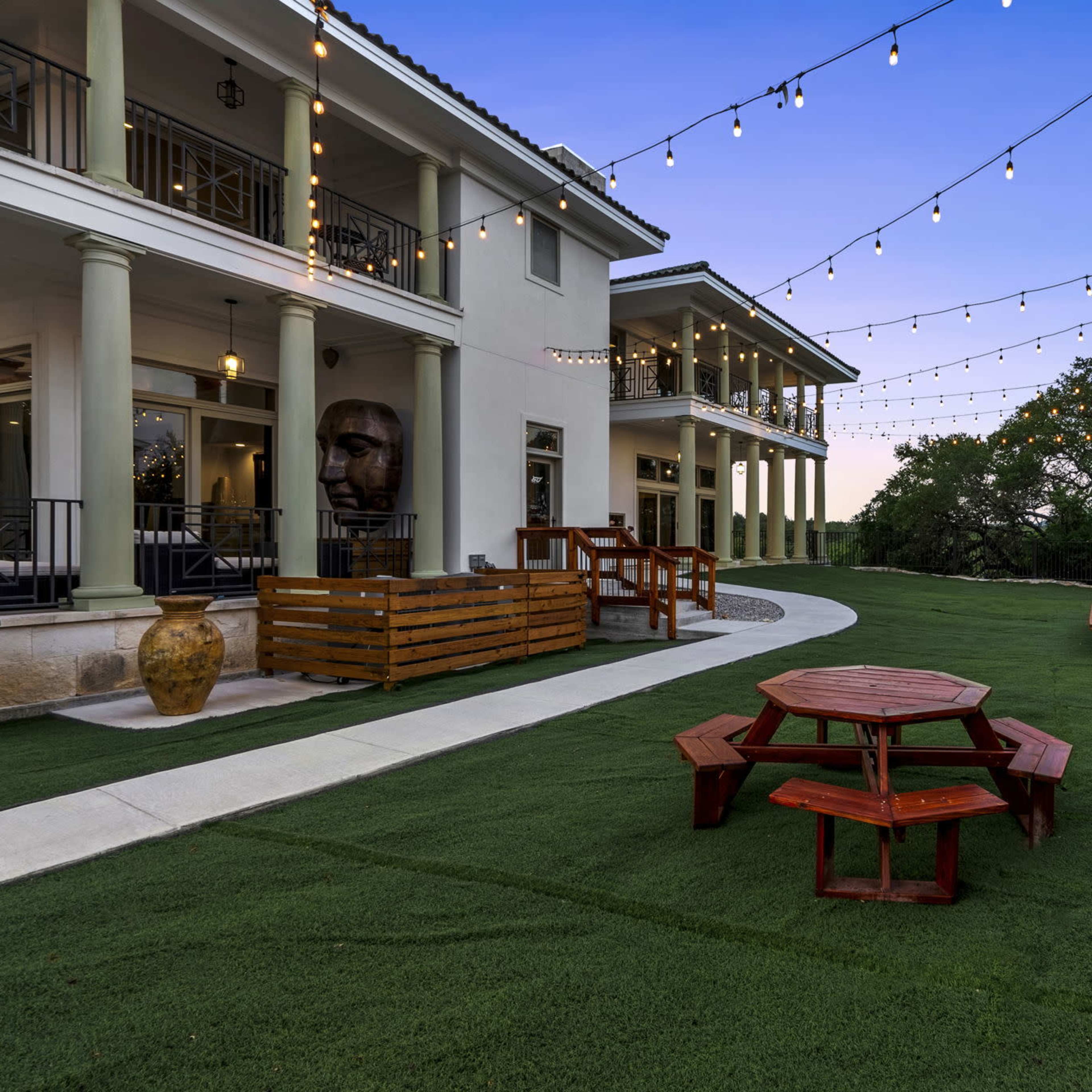 The scene features an outdoor area with string lights overhead, a red picnic table, and a landscaped lawn beside a modern building.