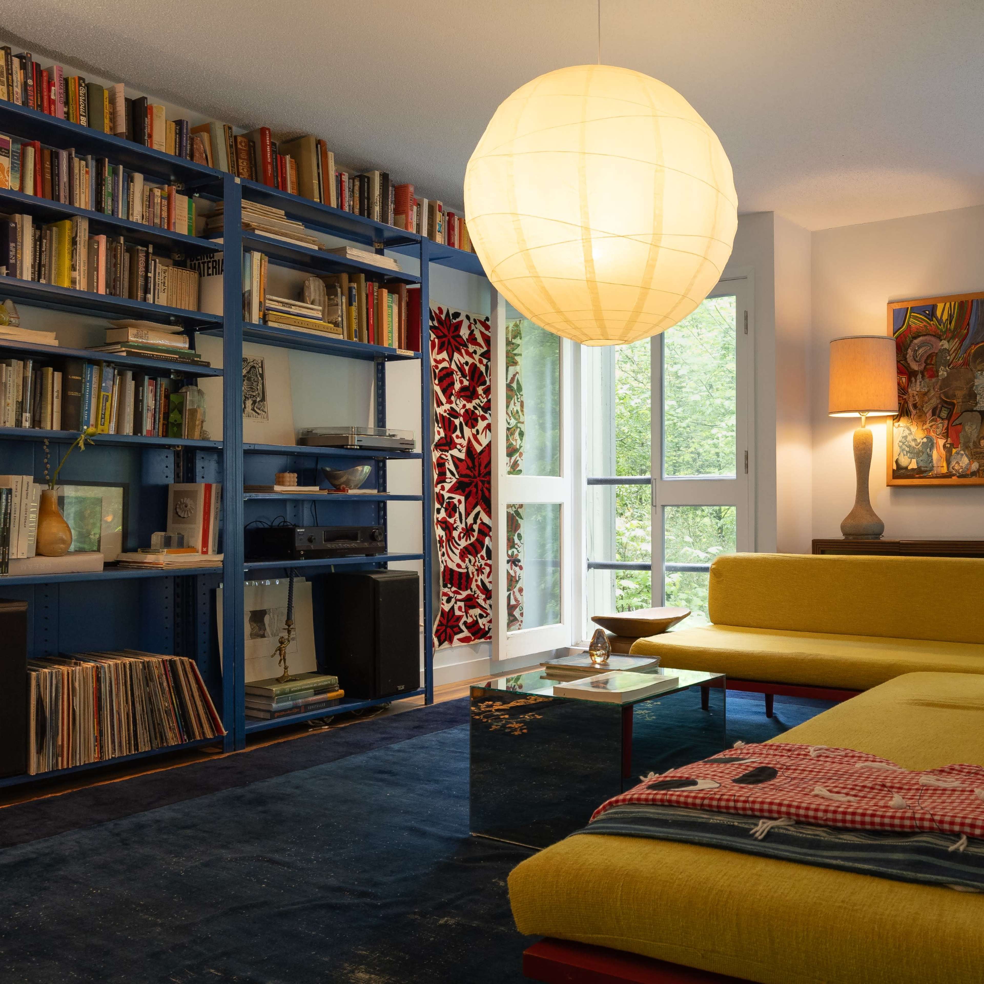 A modern living room featuring a yellow couch, a large round pendant light, bookshelves filled with books, and windows overlooking greenery.
