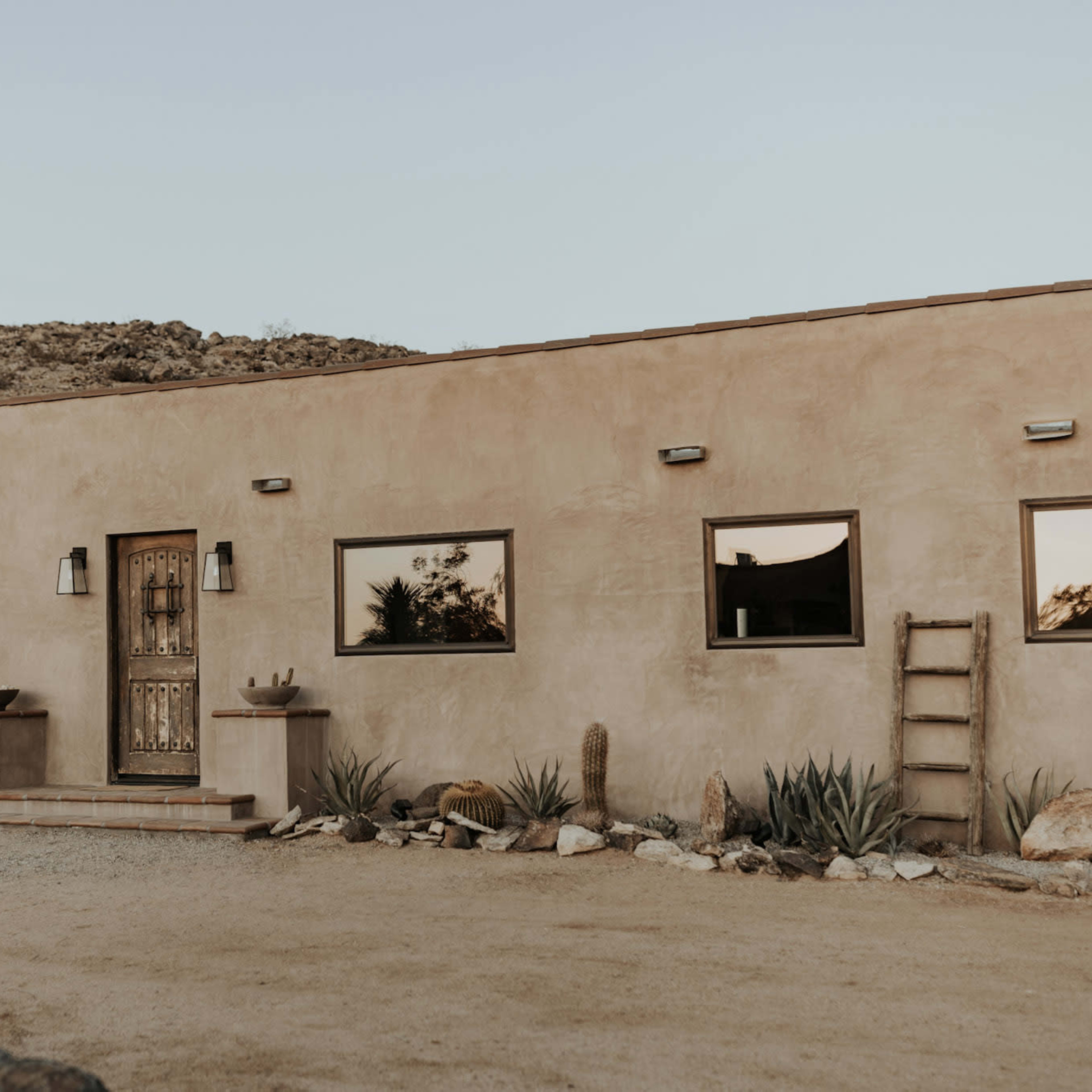 A single-story adobe-style house with a wooden front door and three windows is set in a desert landscape with gravel and various cacti.