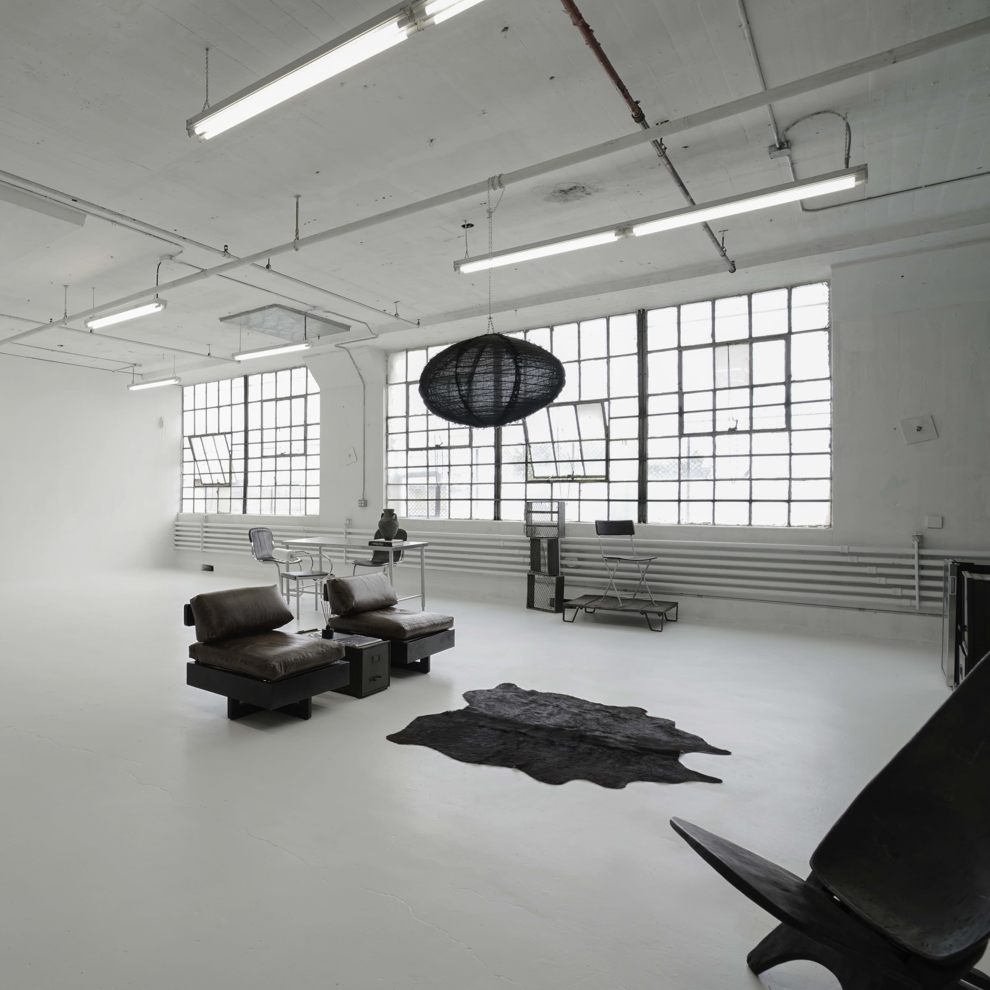 A spacious, minimally furnished room with white walls, large windows, and a few pieces of modern furniture, including two leather chairs and a round pendant light.