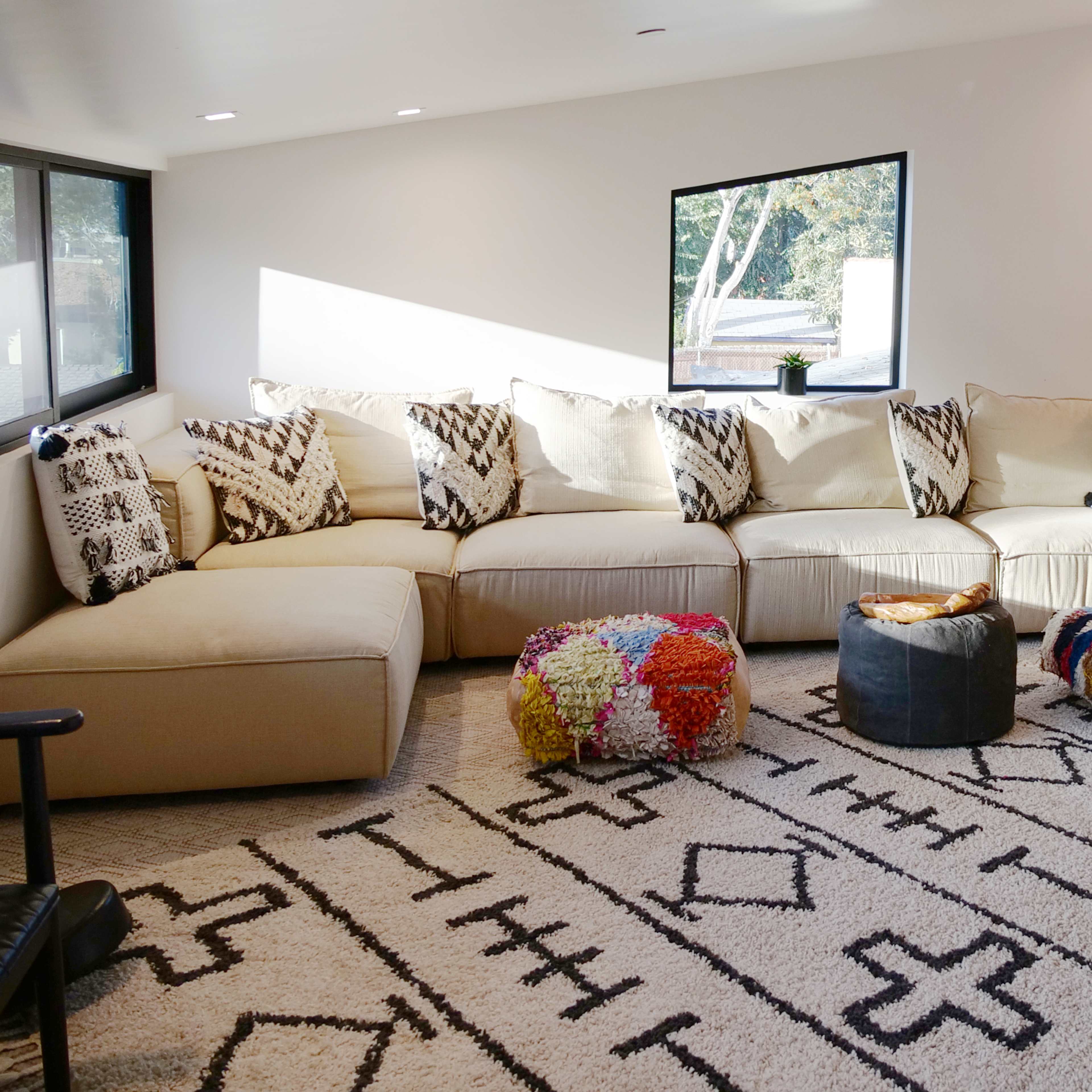 A bright living room with a large sectional couch, a colorful pouf, a patterned rug, and decorative plants.