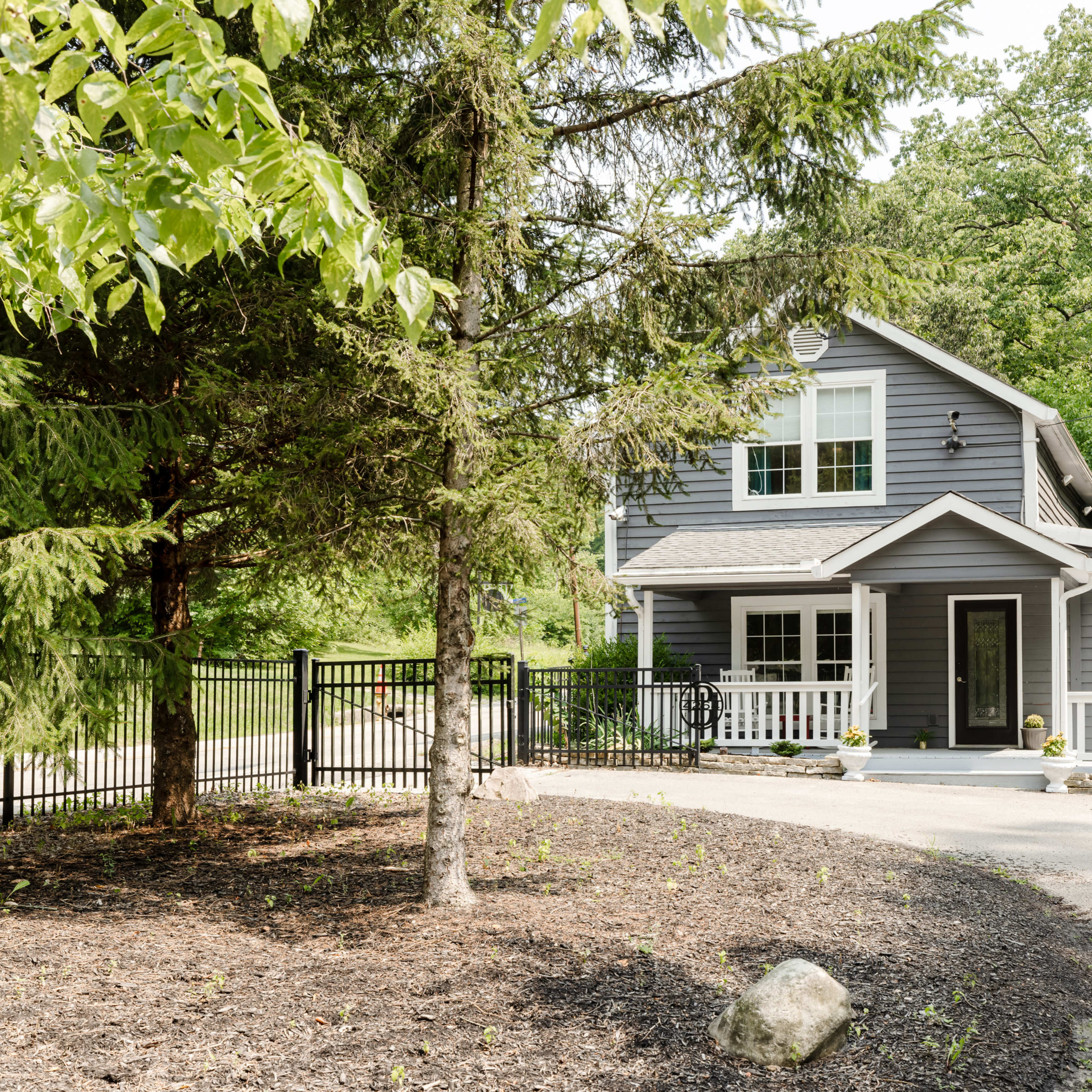 A two-story gray house is situated among trees, with a front porch, white railings, and a circular driveway.
