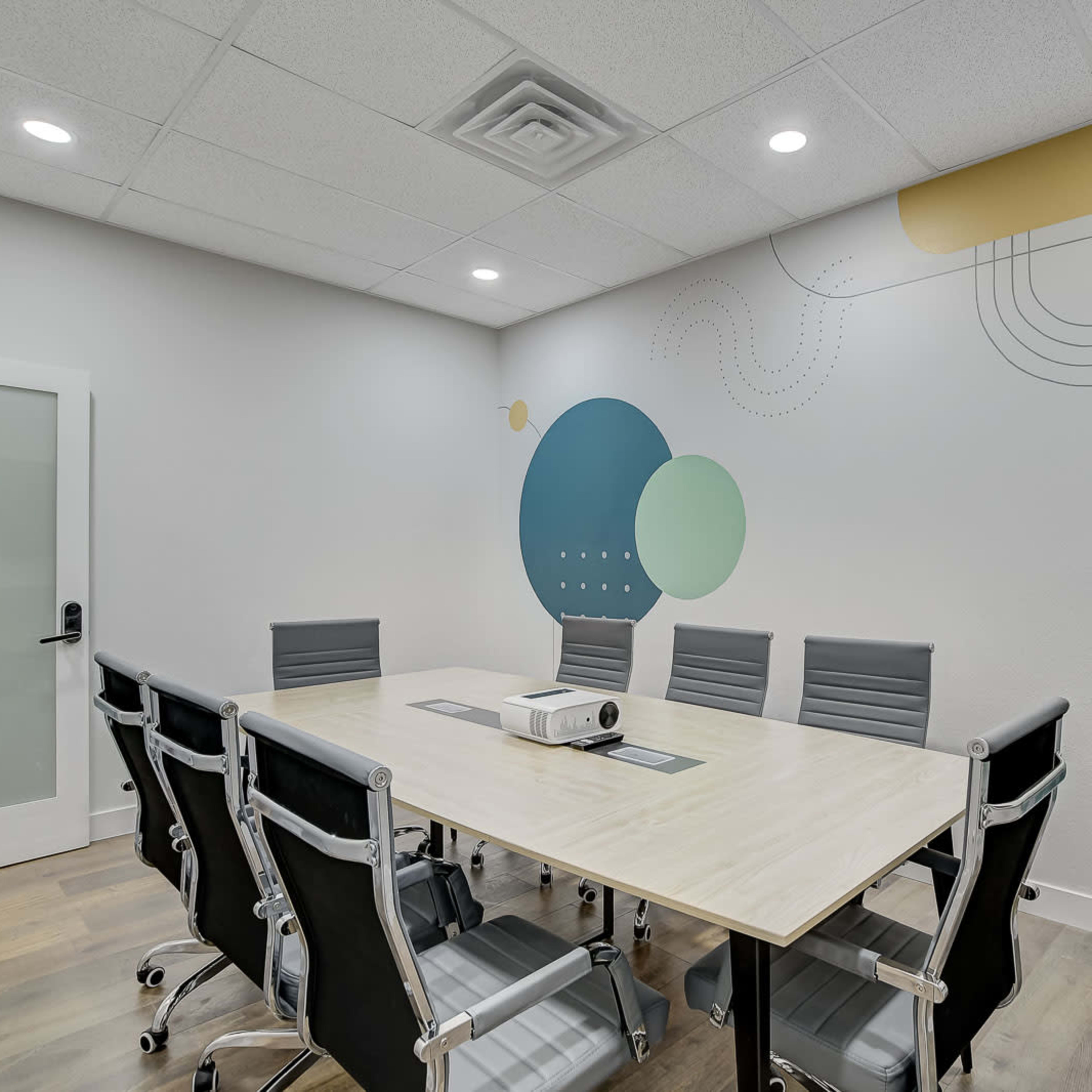 A conference room features a large table surrounded by chairs, with a projector on the table and colorful geometric designs on the wall.