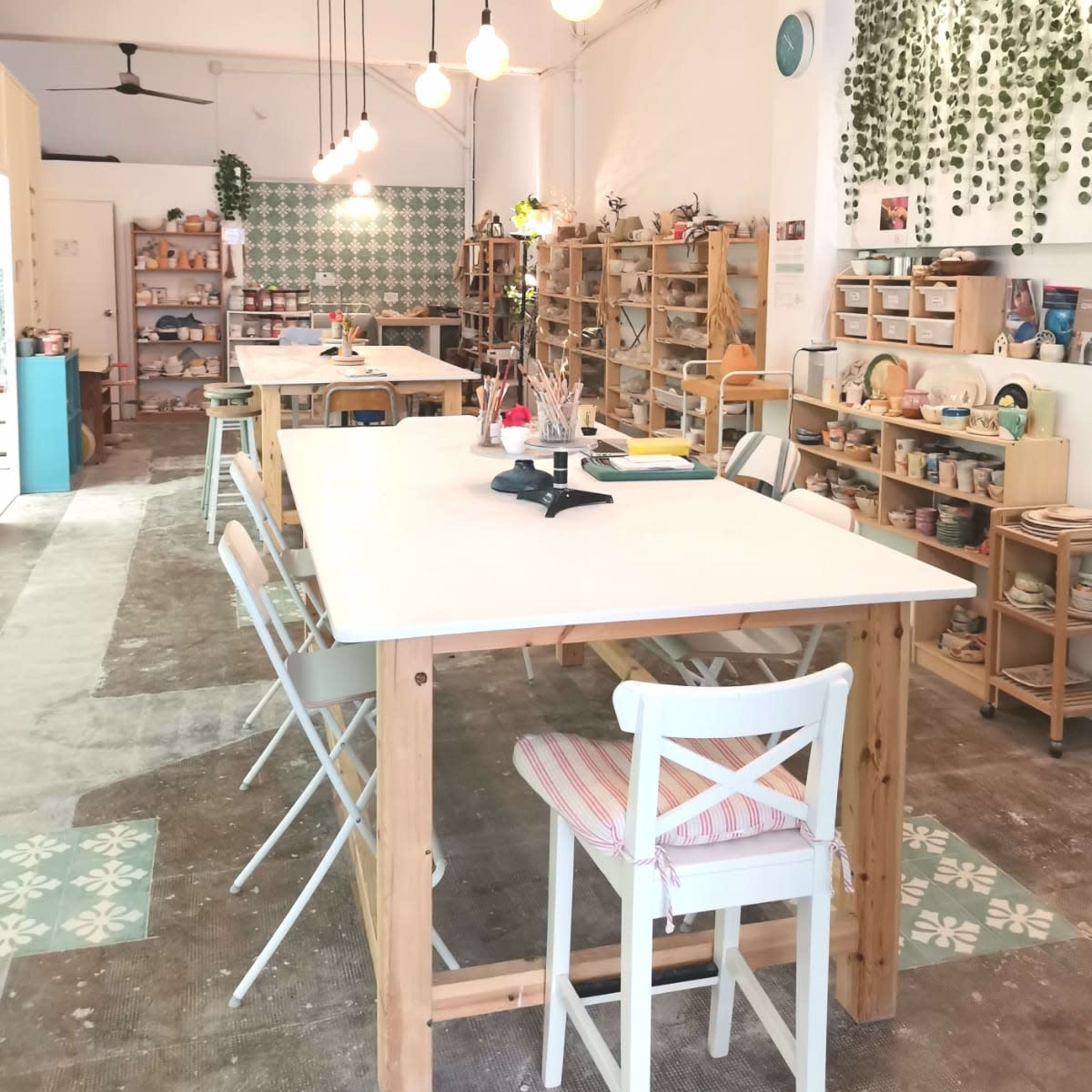 An airy craft studio with wooden tables, shelves filled with art supplies, and pendant lights hanging from the ceiling.