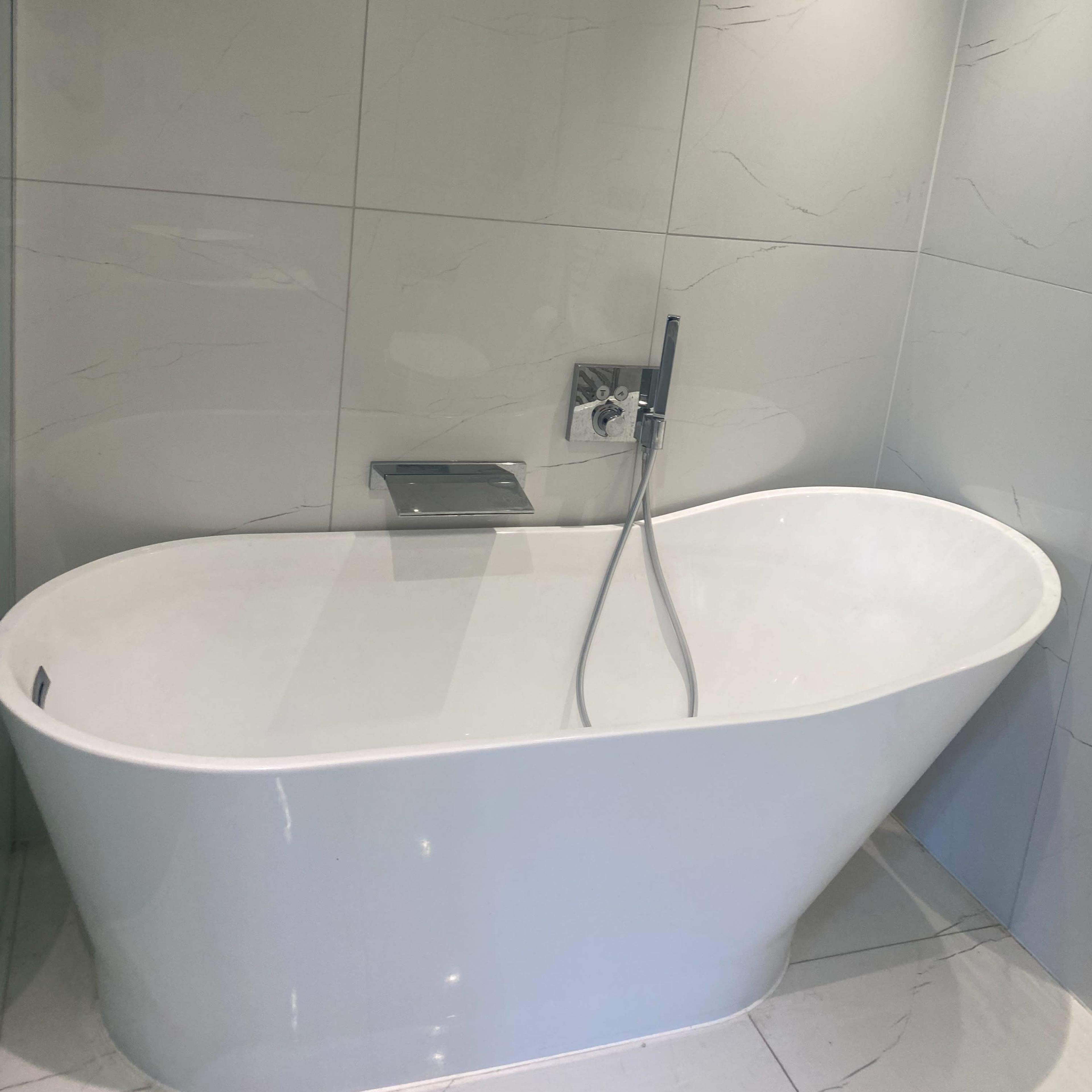 A modern freestanding bathtub is positioned against a marble-tiled wall, featuring a sleek faucet and handheld shower attachment.