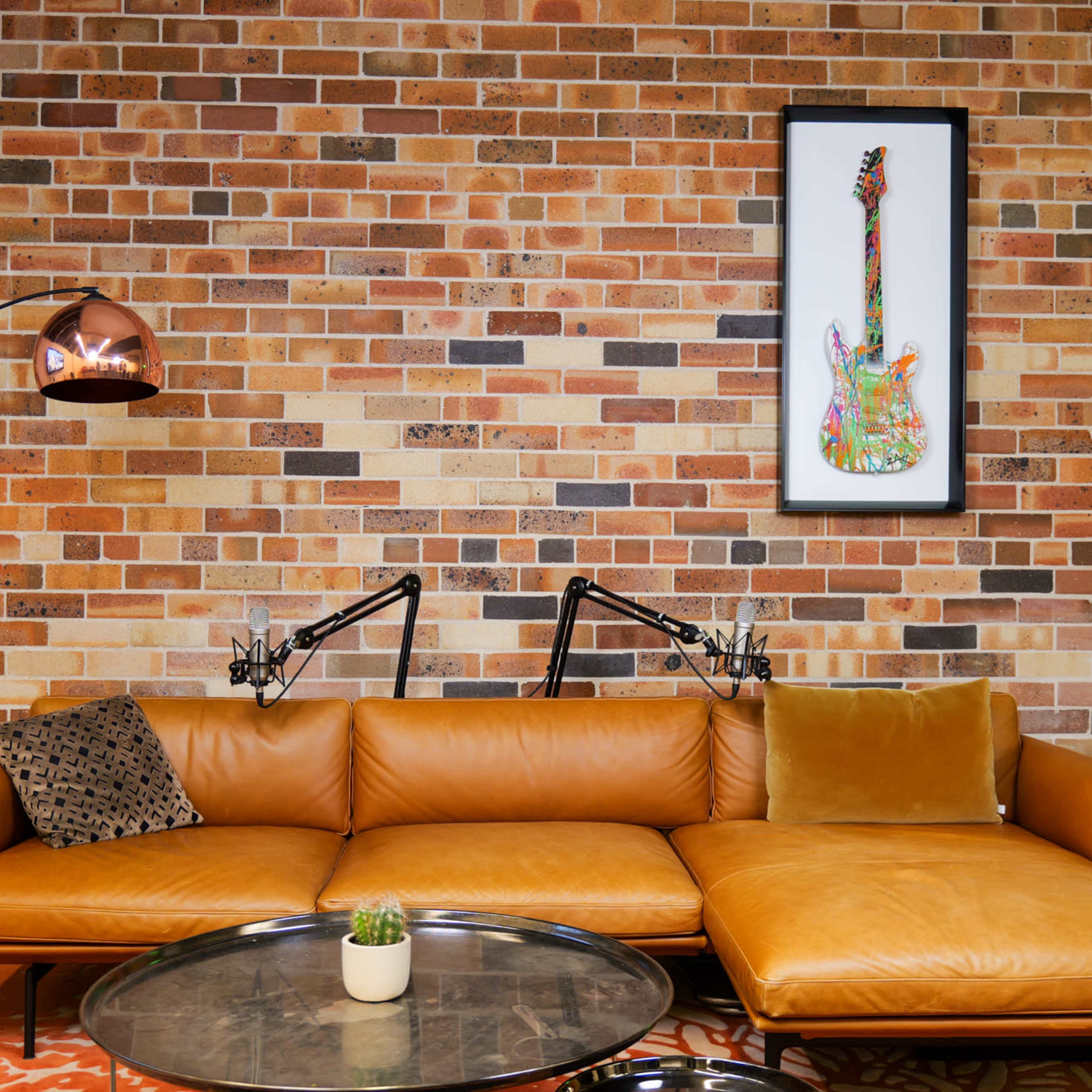 A brown leather sectional sofa is positioned against a brick wall, featuring a framed guitar artwork above and a modern floor lamp beside it.