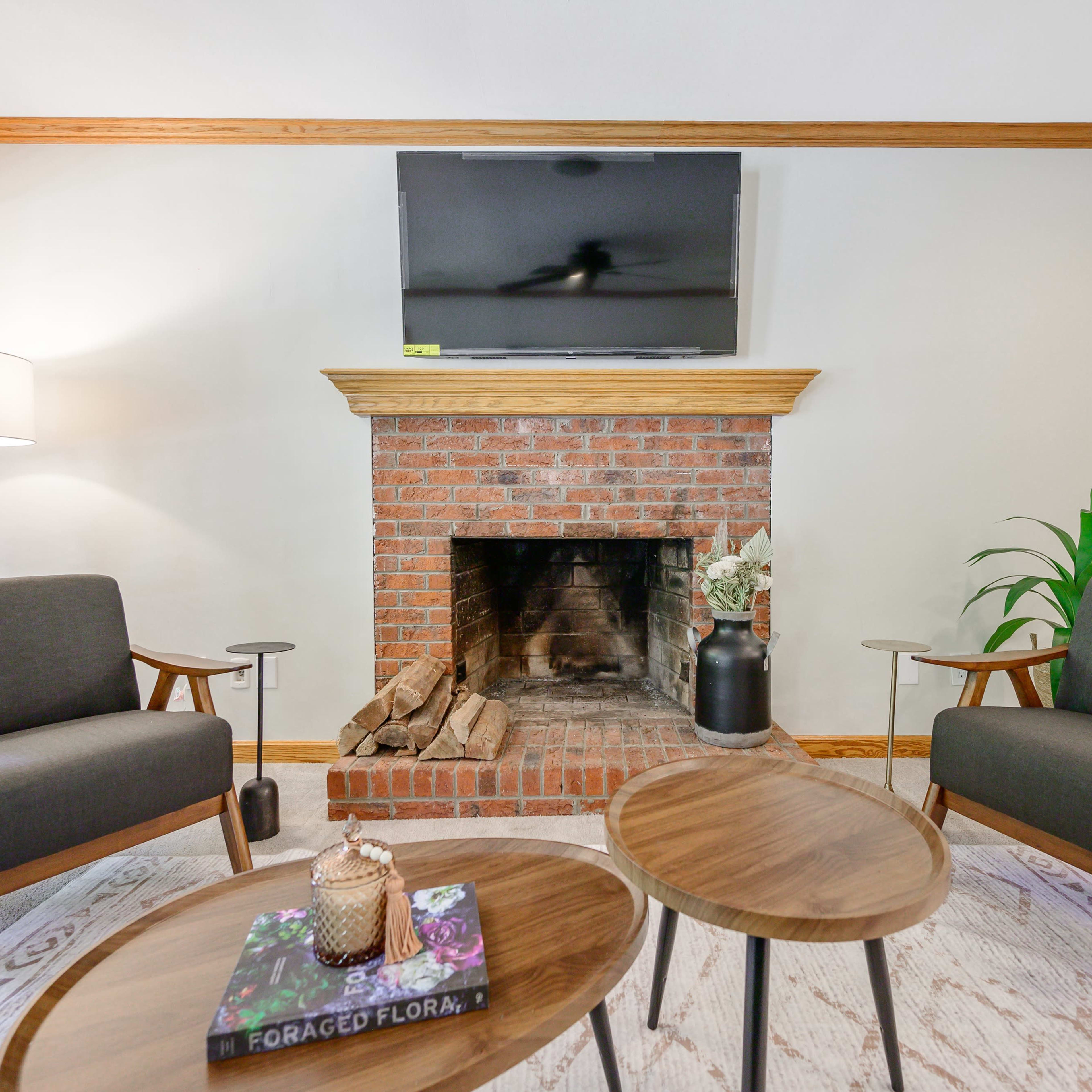 A cozy living room features two gray armchairs facing a brick fireplace with a television mounted above it, accompanied by two wood tables and a floor lamp.