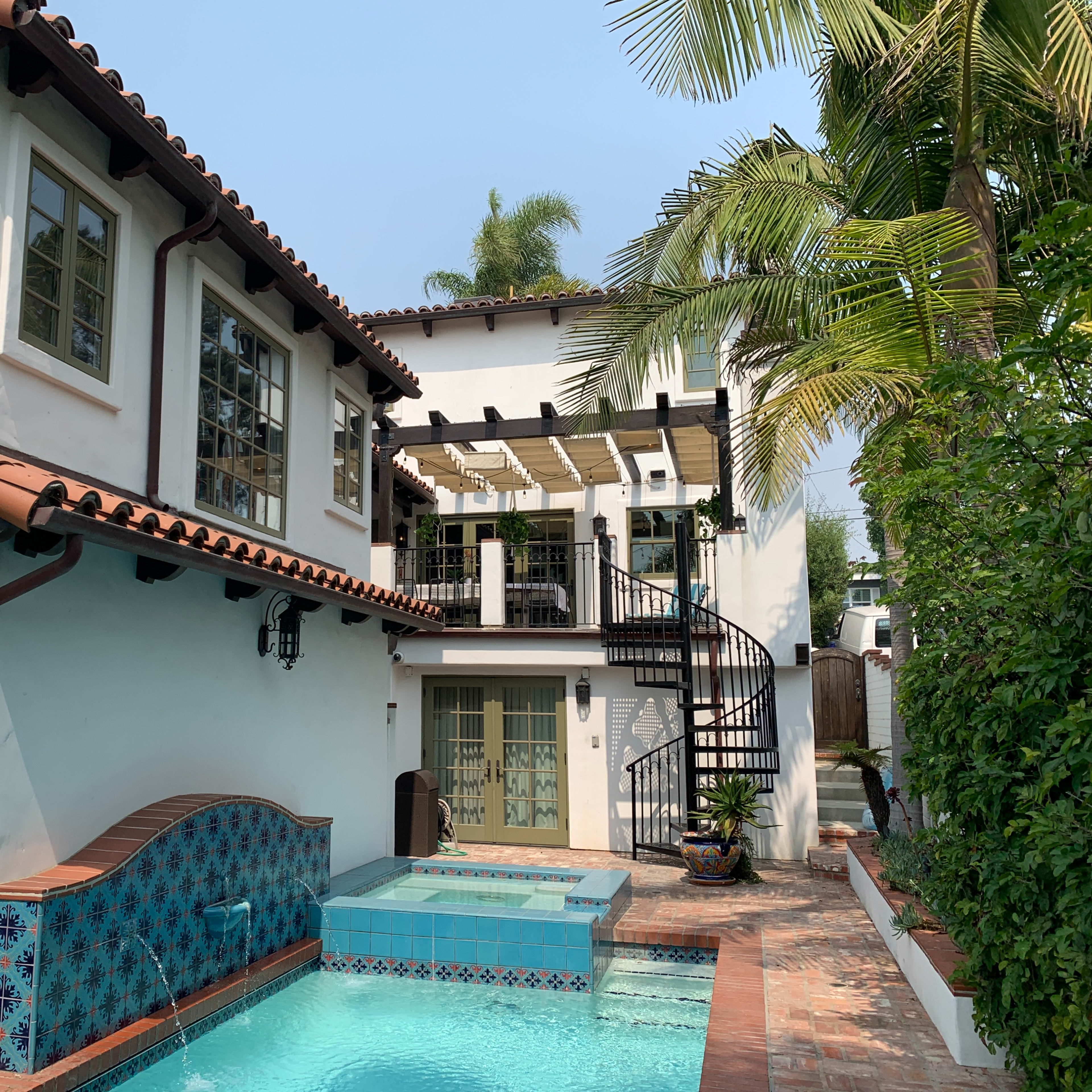 A two-story house with a tiled roof, a small swimming pool, and a spiral staircase leading to a balcony surrounded by palm trees.