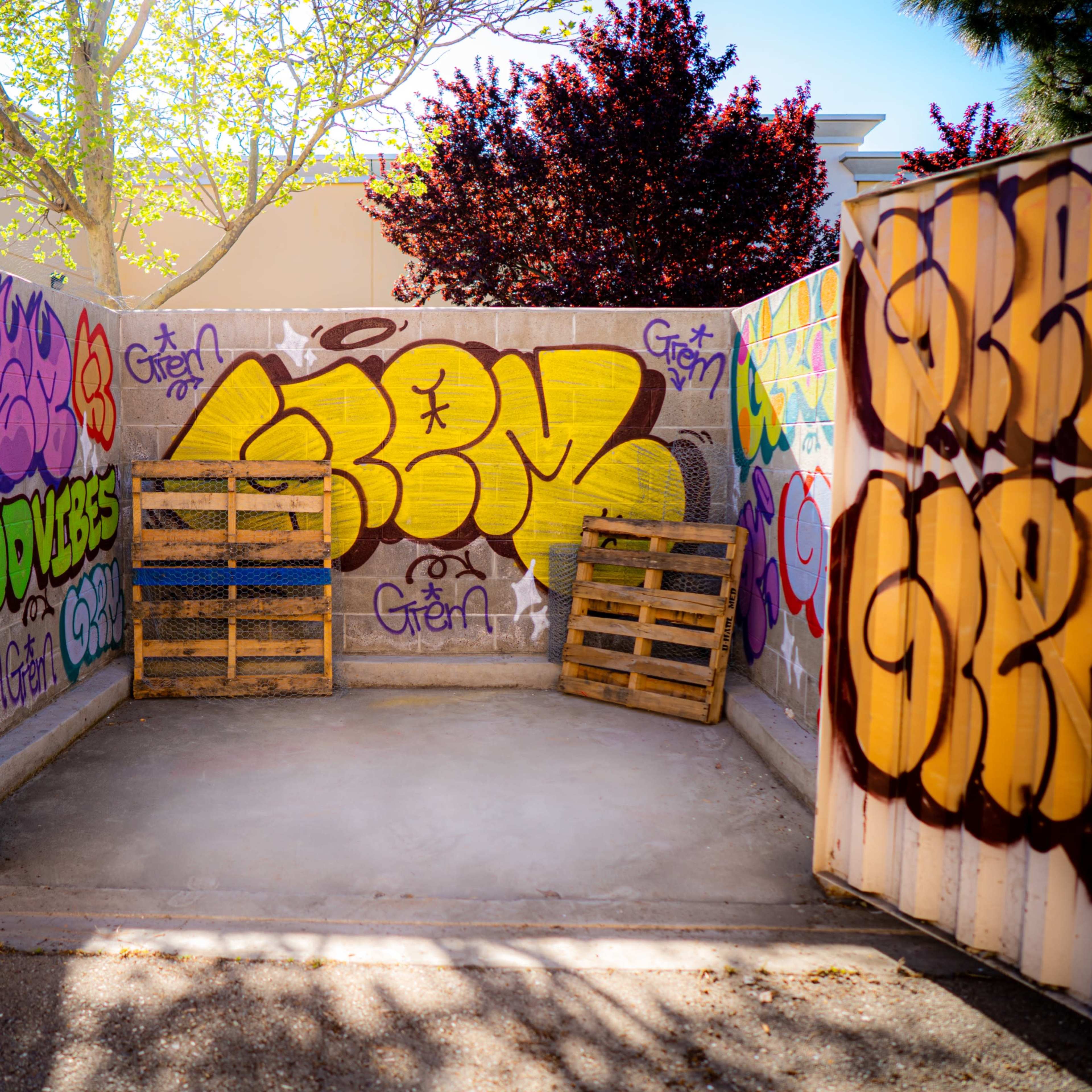 A cement area enclosed by walls covered in colorful graffiti, featuring yellow and purple designs, with wooden pallets stacked inside.