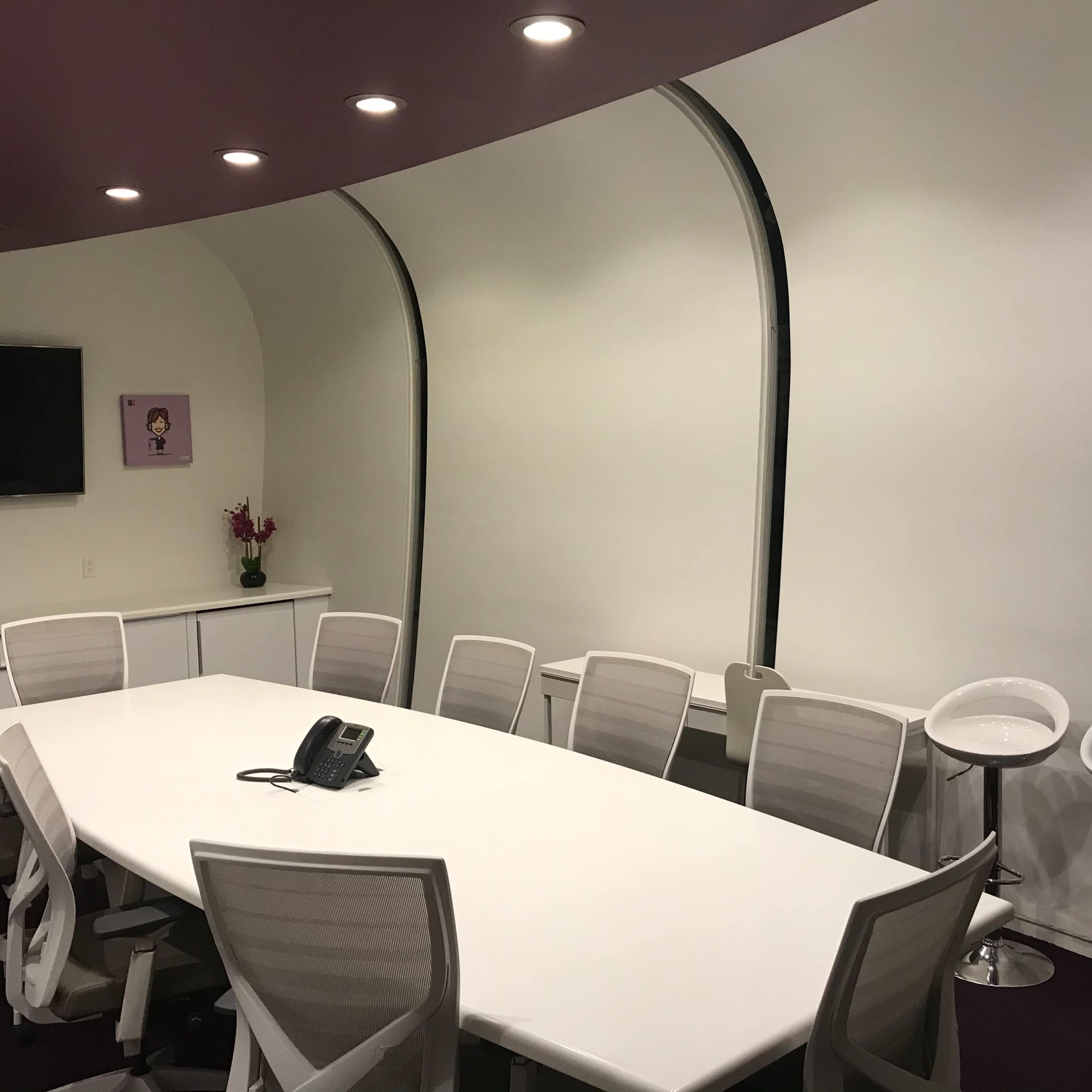 A modern conference room with a large white table, several white chairs, and a wall-mounted television.