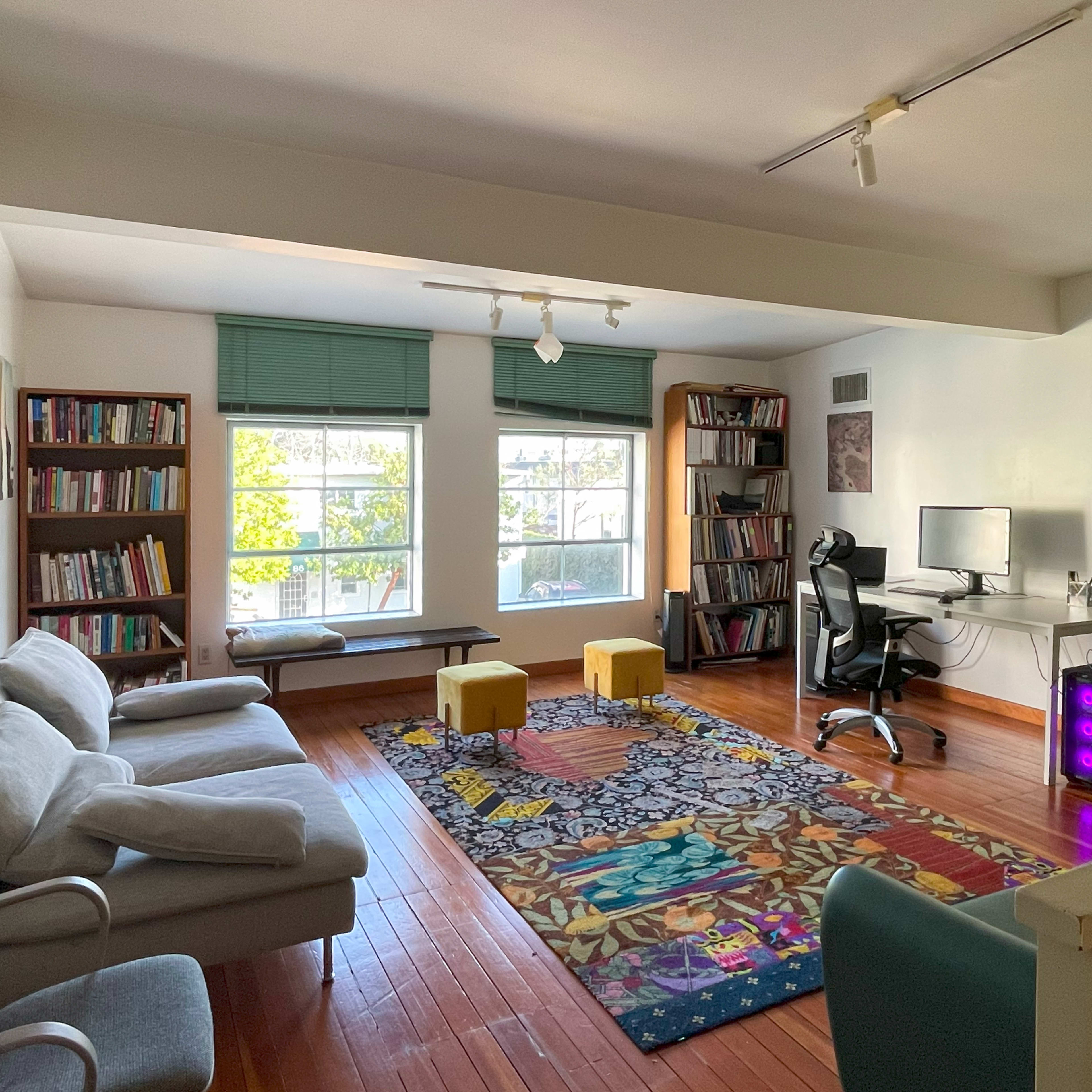 The room features a hardwood floor with a large area rug, a desk with a computer, a bookshelf filled with books, a seating area with two sofas, and two yellow stools.