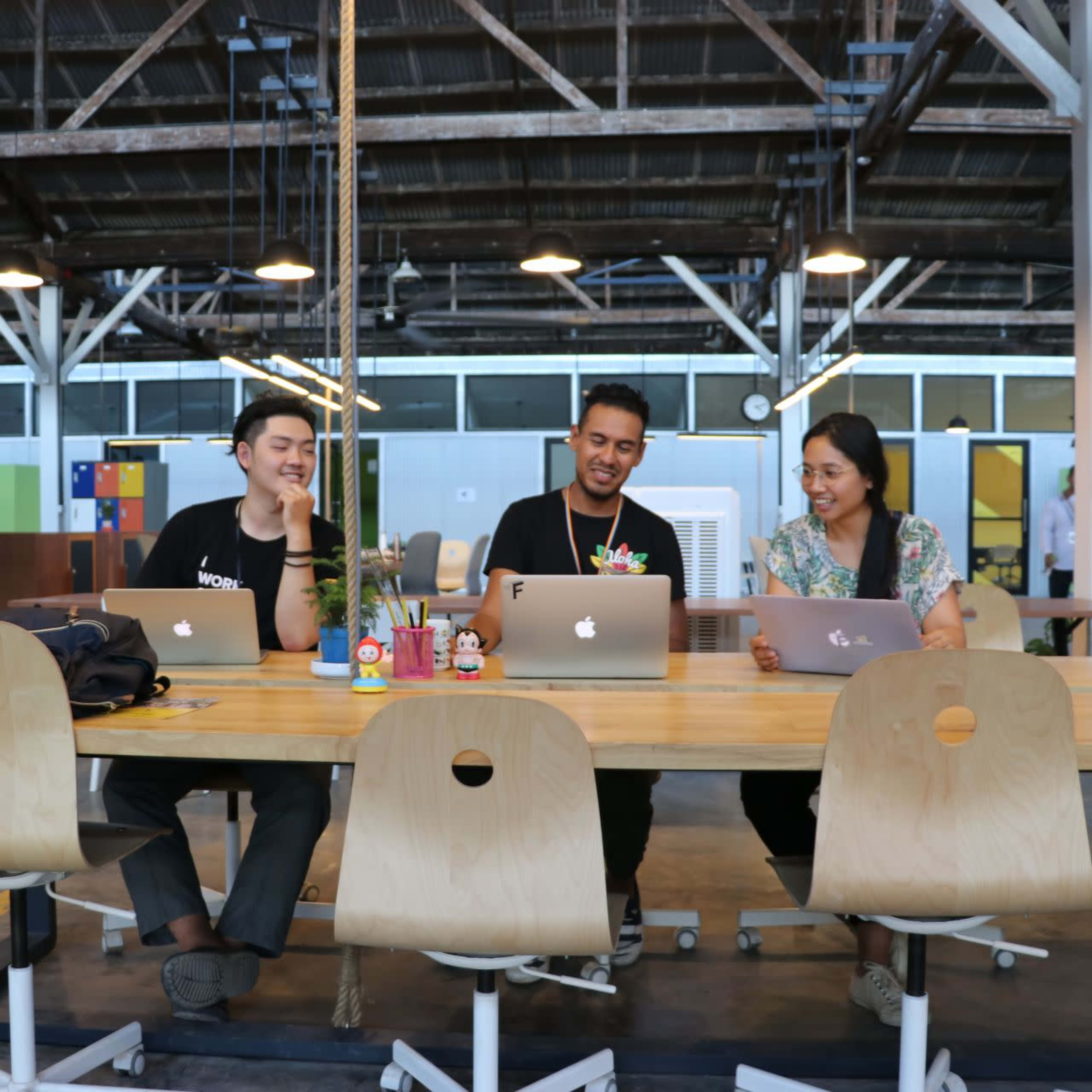 Four people are seated at a wooden table in an open office space, each working on a laptop.
