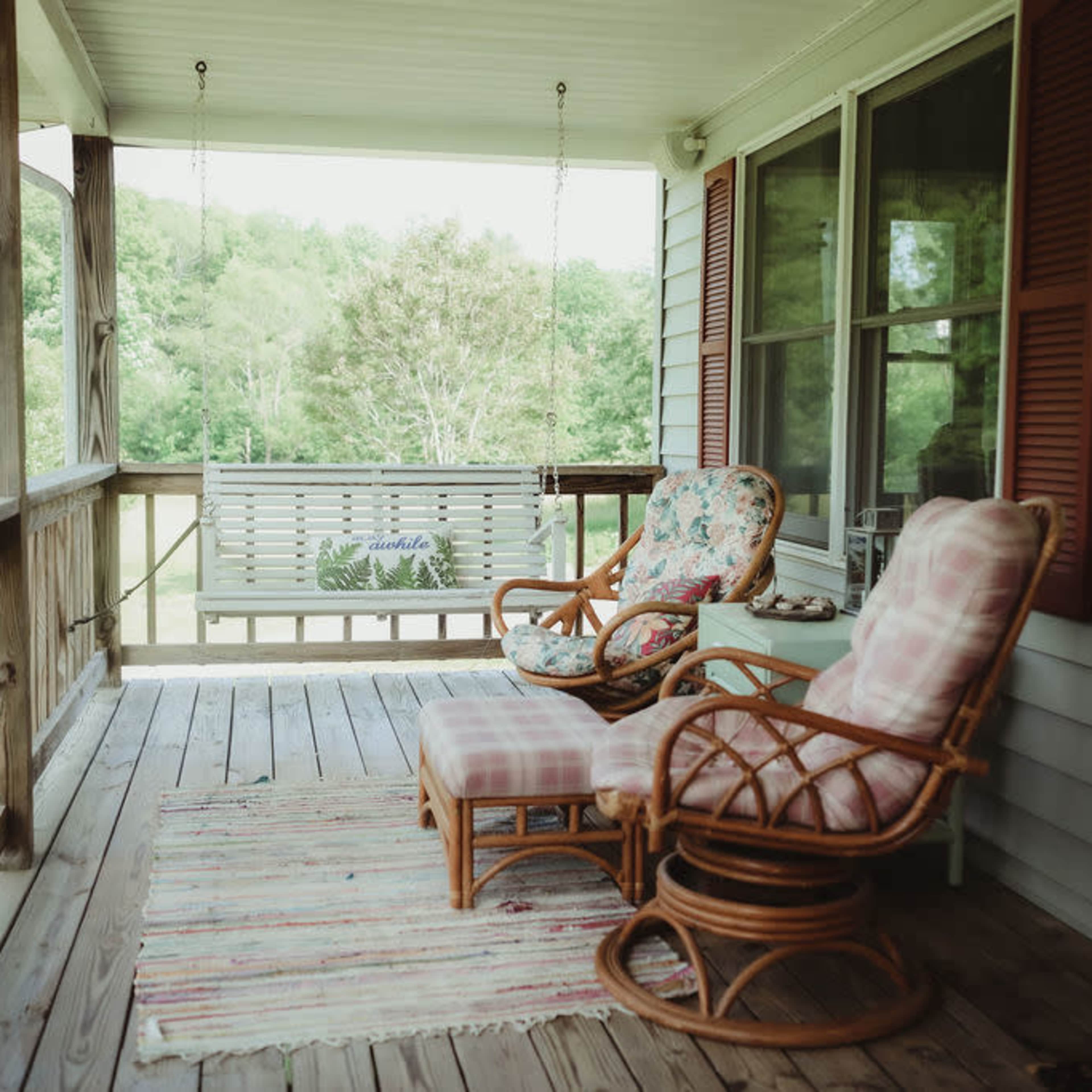 A porch with two wicker chairs, a swing, and a small table, surrounded by greenery.